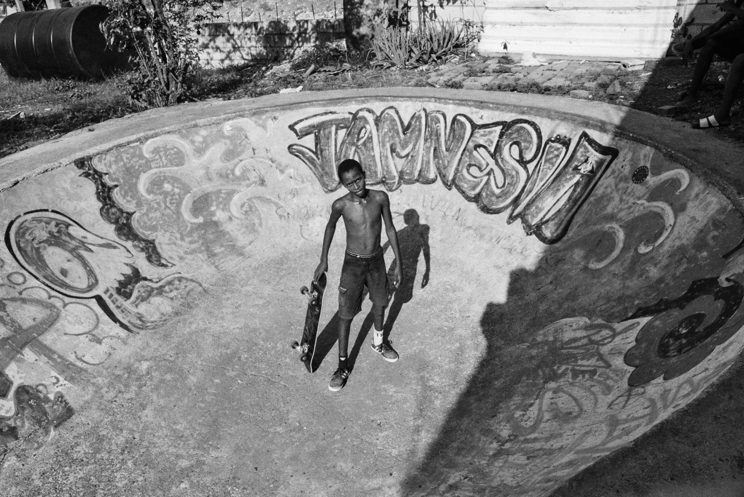 A YOUNG BOY HOLDING A SKATEBOARD STANDING IN FRONT OF A DECORATED CONCRETE BOWL AT A SKATEPARK, WITH A GRAFFITI DESIGN AND THE WORD 'VAMOS' VISIBLE ON THE WALL.