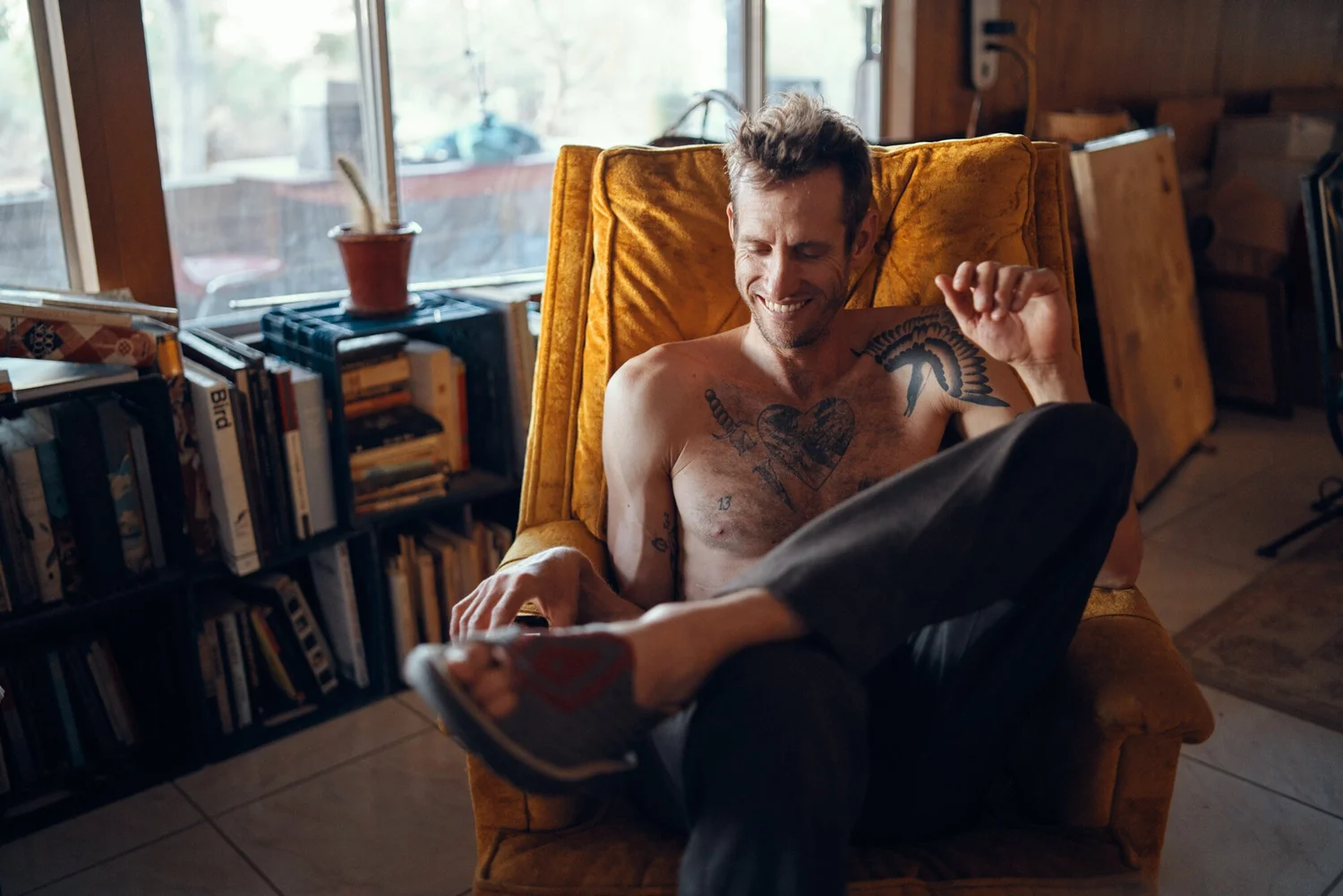 A SHIRTLESS MAN WITH TATTOOS SMILING AND READING A BOOK IN A COZY ROOM WITH A YELLOW ARMCHAIR, A BOOKSHELF, AND A LARGE WINDOW.