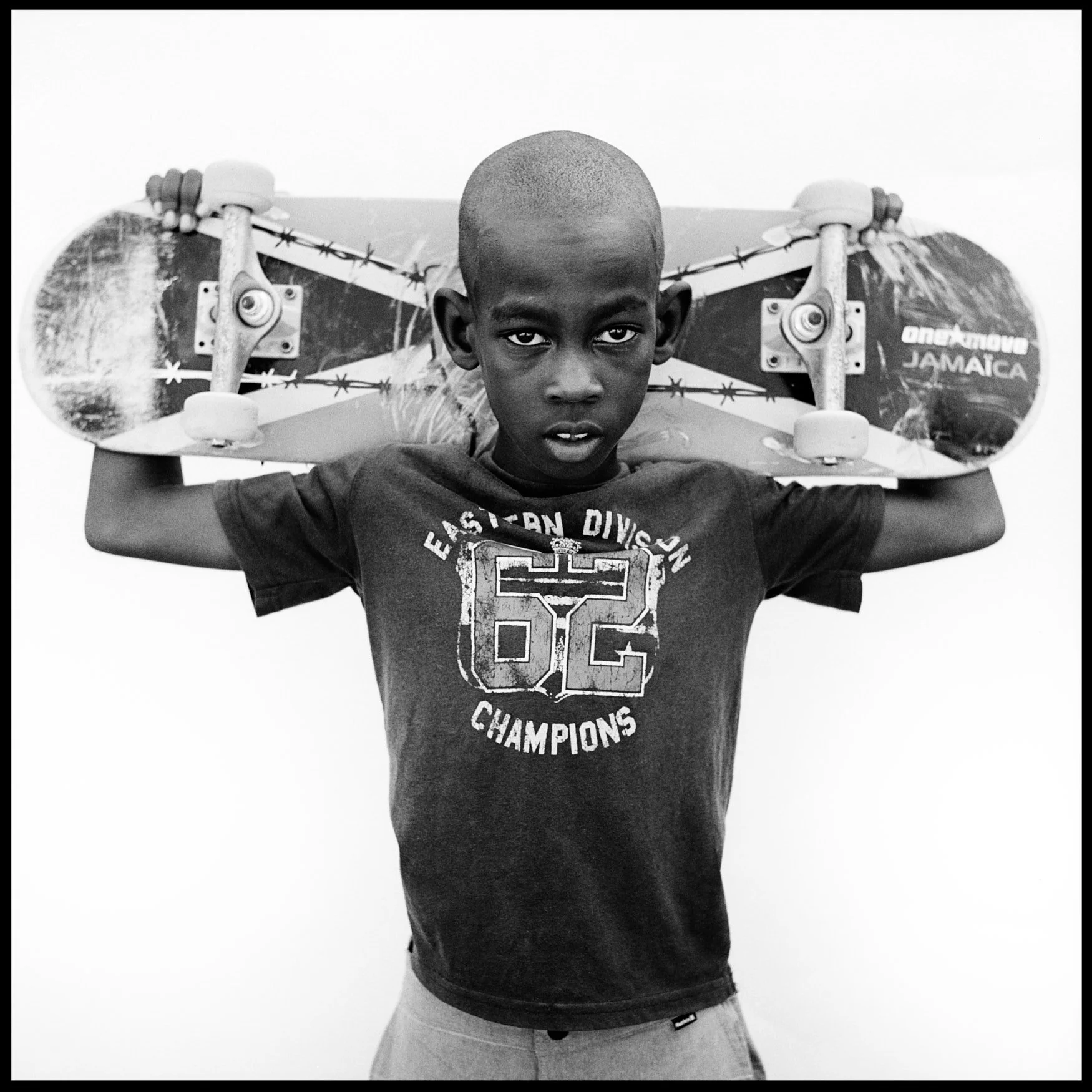 A JAMAICAN BOY HOLDING A SKATEBOARD ACROSS HIS SHOULDERS AND LOOKING AT THE CAMERA, BLACK-AND-WHITE PORTRAIT.