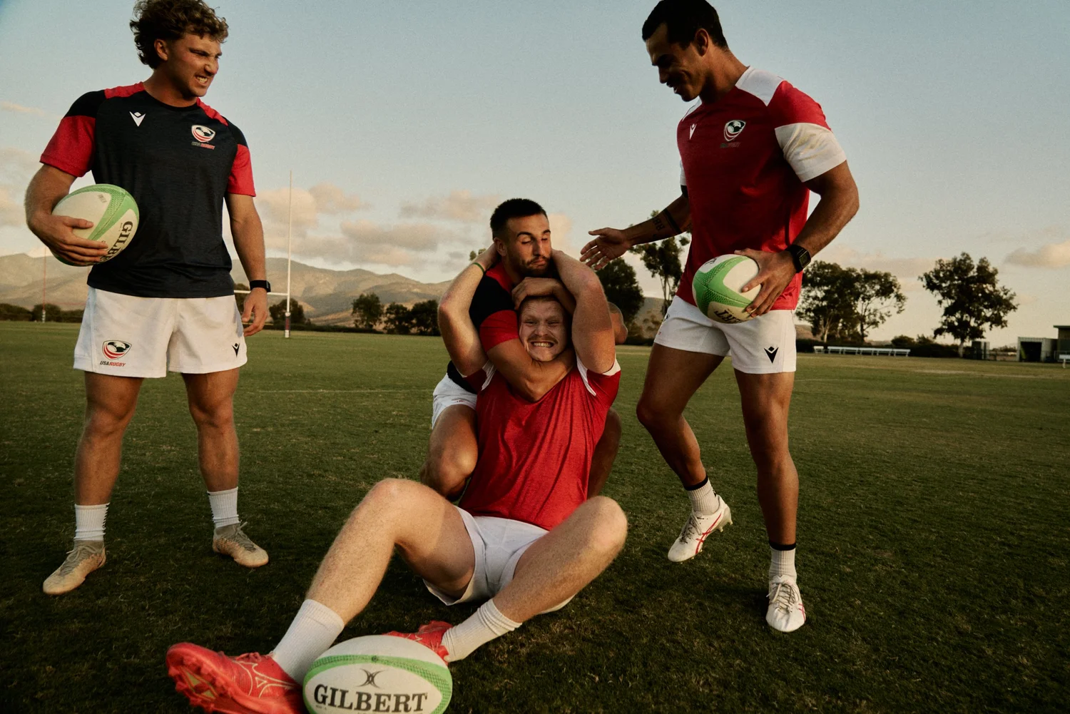 FOUR RUGBY PLAYERS ON A FIELD DURING SUNSET, WITH THREE STANDING AND ONE SITTING ON THE GROUND. THE PLAYERS ARE WEARING RED AND BLACK UNIFORMS WITH RUGBY BALLS, AND A SCENIC MOUNTAIN LANDSCAPE IN THE BACKGROUND.