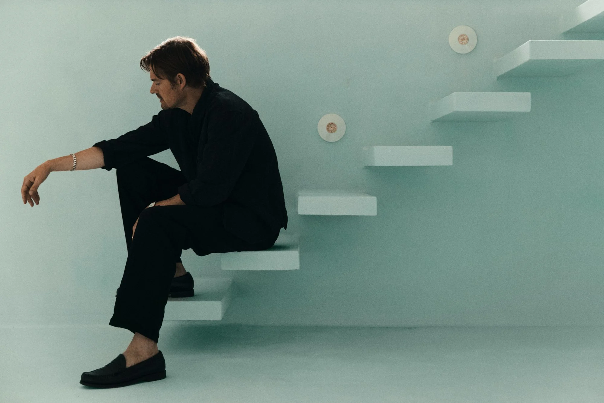 ACTOR SAM RILEY SEATED ON WHITE STAIRS AGAINST A PALE TURQUOISE WALL, DRESSED IN BLACK.