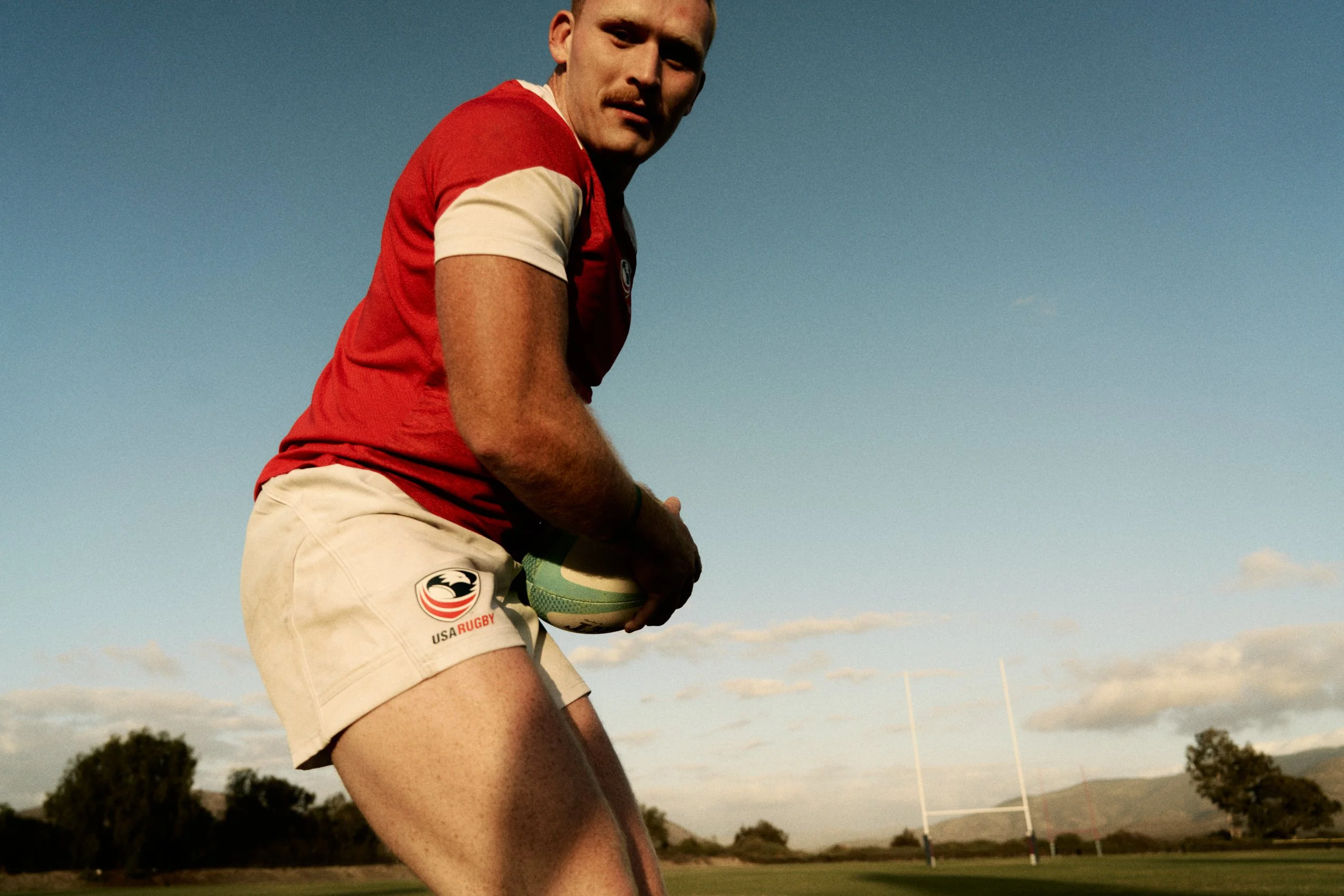 A MAN IN A RED SHIRT AND BEIGE SHORTS HOLDING A RUGBY BALL DURING DAYTIME OUTDOORS WITH A RUGBY FIELD AND GOALPOSTS IN THE BACKGROUND.