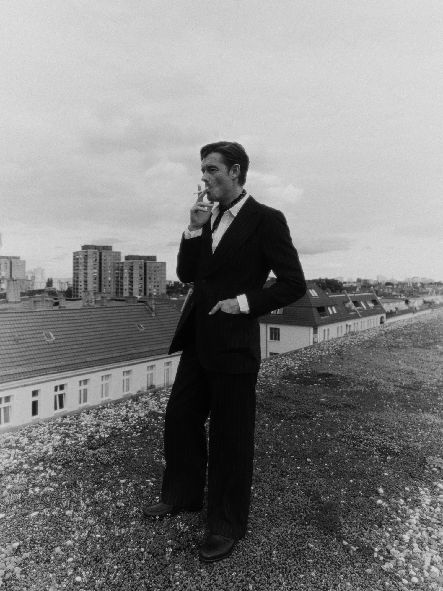 ACTOR SAM RILEY SMOKING A CIGARETTE IN A SUIT ON A BERLIN ROOFTOP, BLACK-AND-WHITE PORTRAIT.