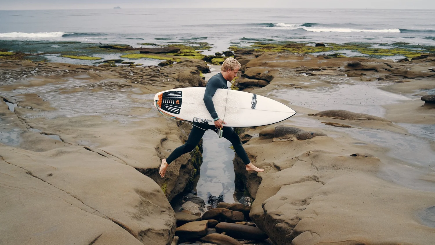 A MAN CARRYING A SURFBOARD WALKING BAREFOOT ON ROCKS NEAR THE OCEAN WITH WAVES AND MOSS-COVERED ROCKS IN THE BACKGROUND.