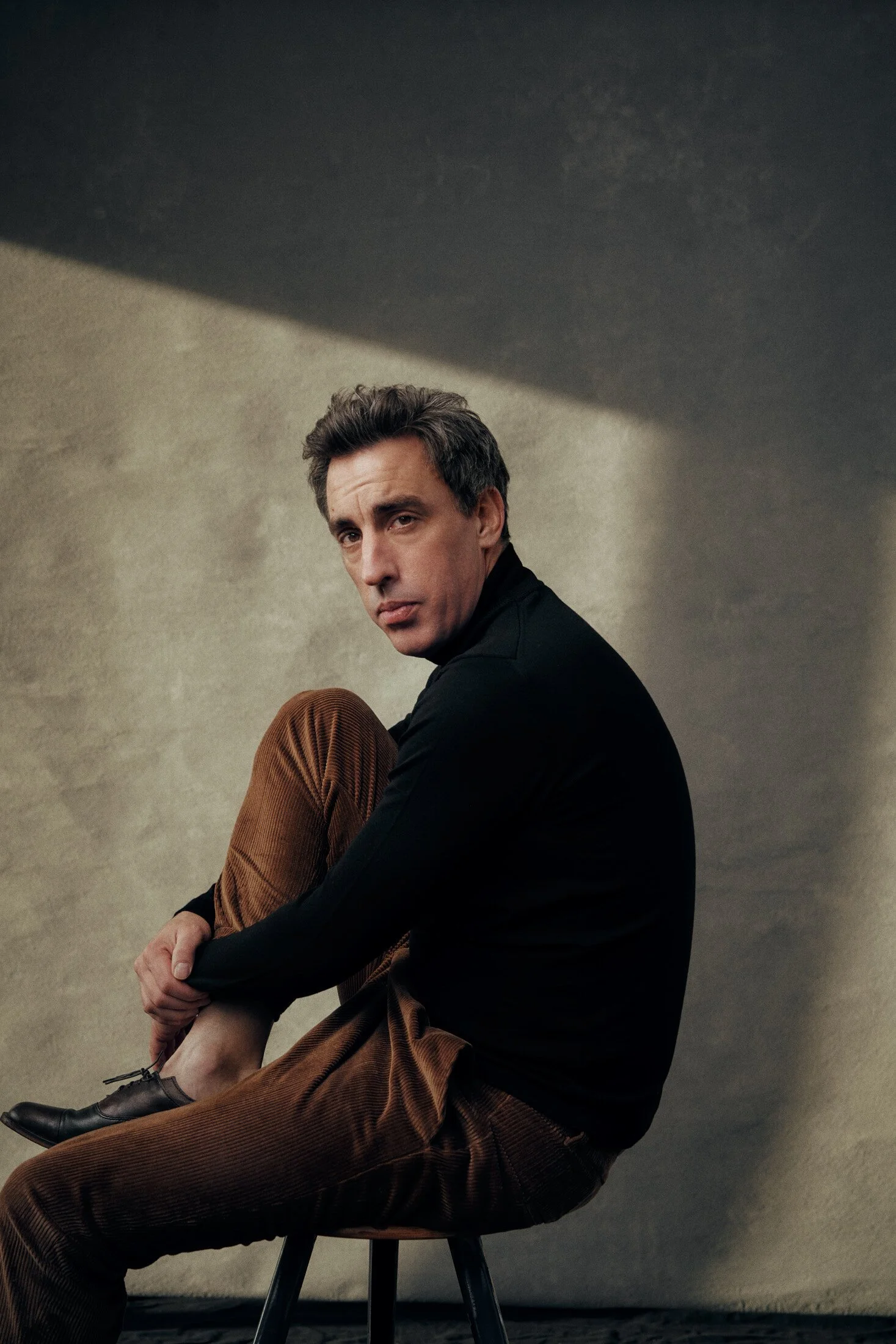 ACTOR ALEXANDER SCHUBERT IS SITTING ON A BLACK CHAIR AGAINST A PLAIN GRAY WALL, WEARING A BLACK SHIRT AND BROWN CORDUROY PANTS, LOOKING THOUGHTFULLY AT THE CAMERA WITH ONE ARM RESTING ON HIS KNEE.