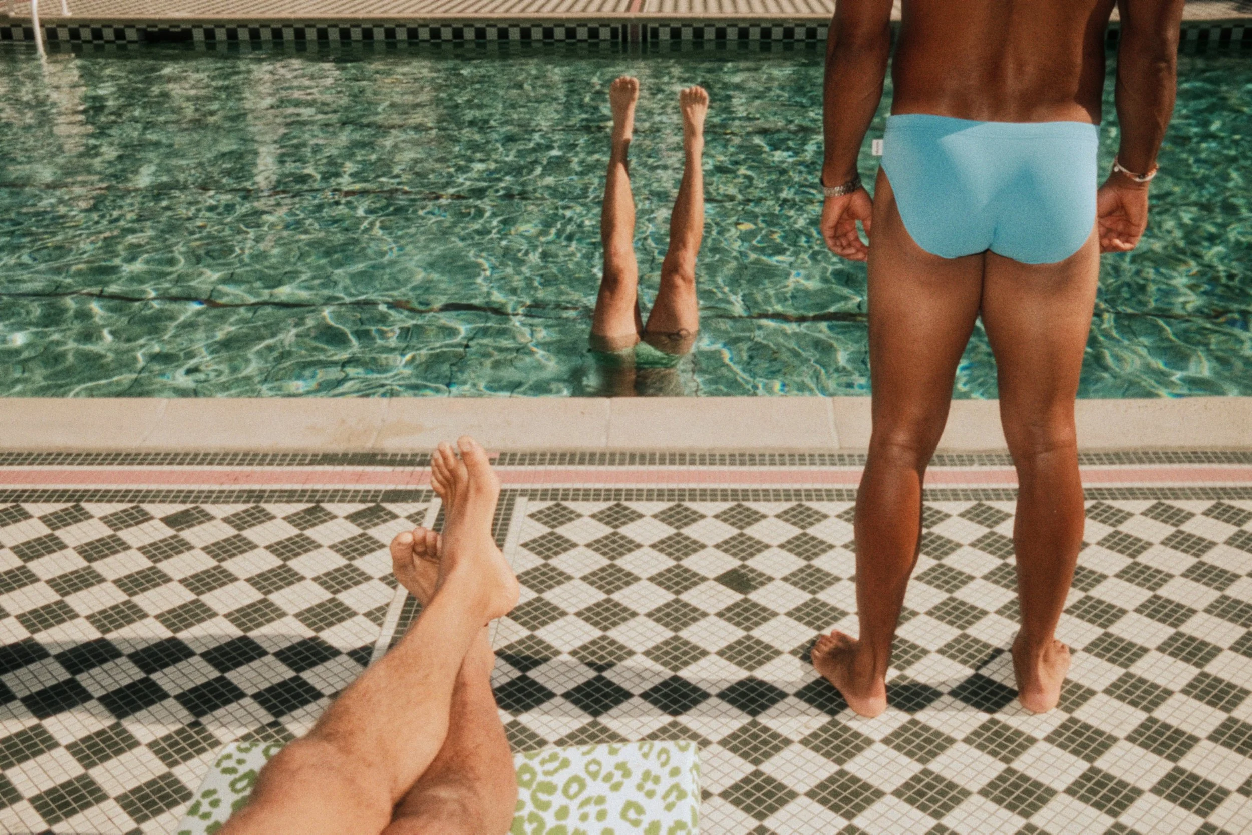 THREE SETS OF LEGS AT A SWIMMING POOL, WITH ONE PAIR STICKING OUT OF THE WATER, FROM NOODLE BAGZ.
