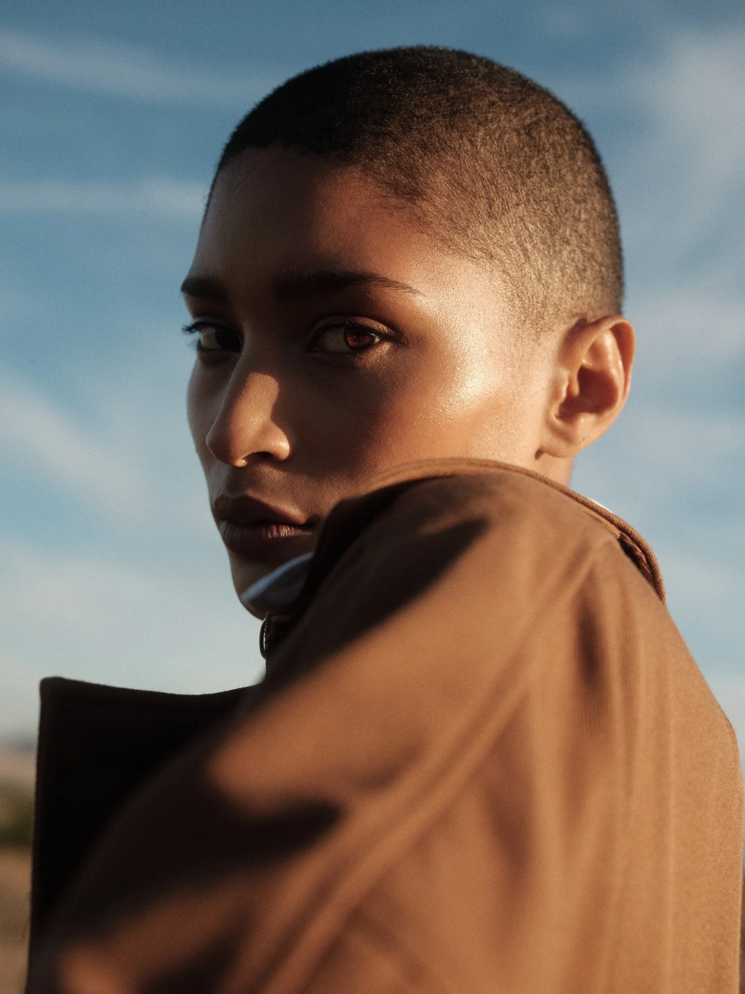 CLOSE-UP PORTRAIT OF ANTONIA LOOKING OVER HER SHOULDER IN A BROWN JACKET AGAINST A BLUE SKY.