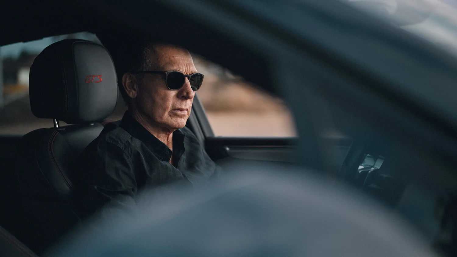 A MAN WEARING BLACK SUNGLASSES SEATED IN A PORSCHE, WITH GTS EMBROIDERED ON THE HEADREST.