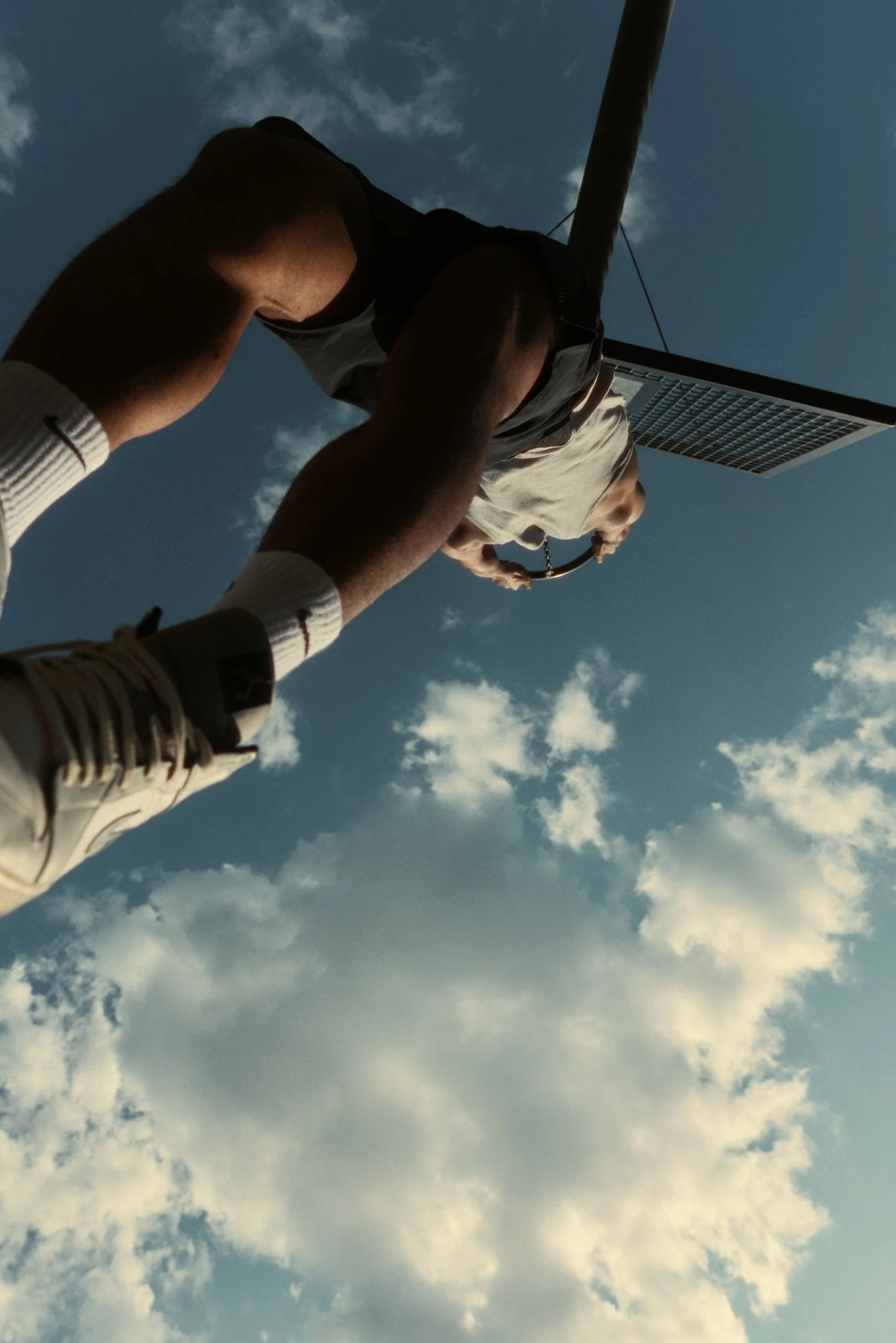 A BASKETBALL PLAYER HANGING FROM THE RIM, PHOTOGRAPHED FROM DIRECTLY BELOW AGAINST A BLUE SKY.