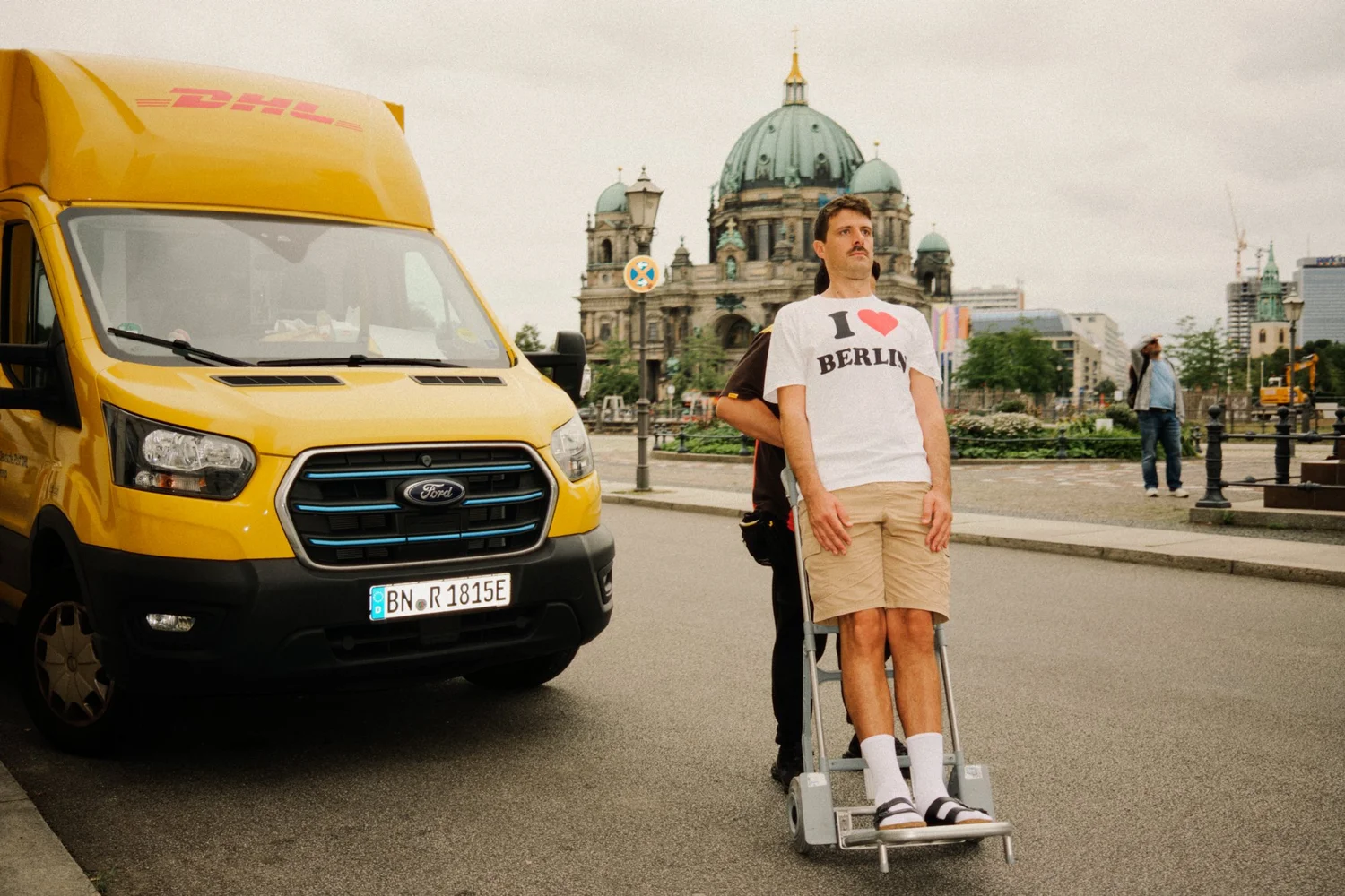A PERSON WITH A MUSTACHE WEARING A WHITE "I LOVE BERLIN" T-SHIRT AND KHAKI SHORTS, USING CRUTCHES, STANDS ON A CITY STREET NEXT TO A BRIGHT YELLOW DHL DELIVERY VAN. IN THE BACKGROUND, AN ORNATE HISTORICAL BUILDING AND A FEW PEDESTRIANS ARE VISIBLE.