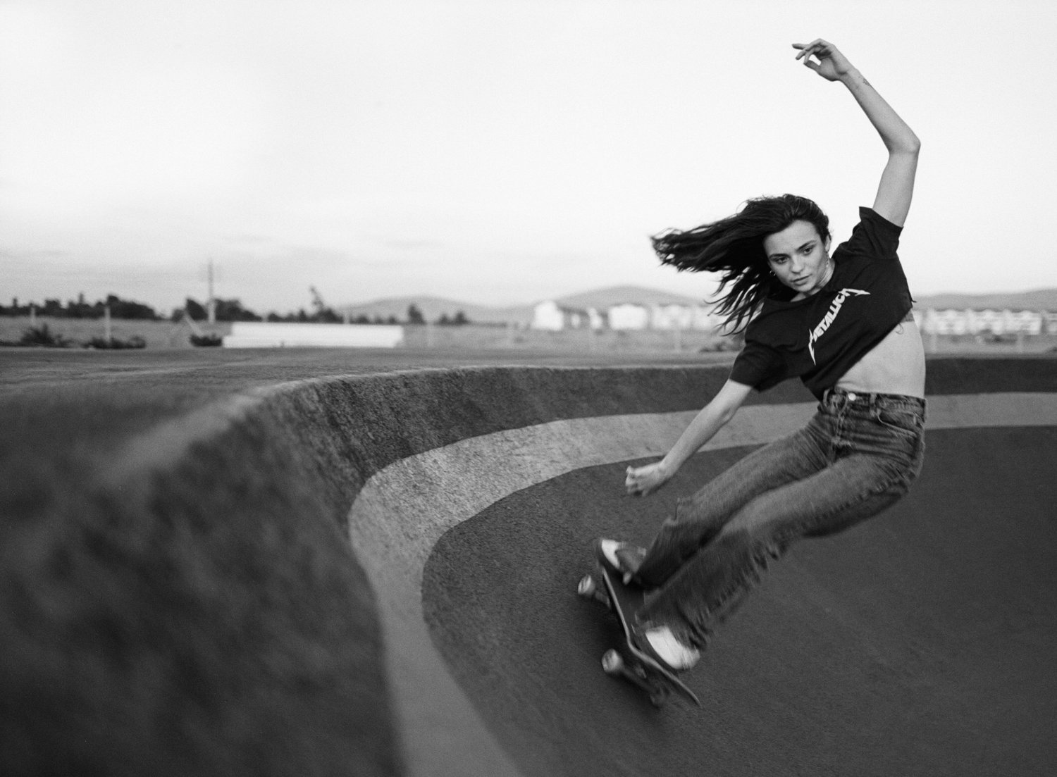 BLACK-AND-WHITE PHOTOGRAPH OF MARIE SKATEBOARDING ON A CURVED CONCRETE RAMP OUTDOORS, WITH HER HAIR FLOWING AND ONE ARM RAISED.