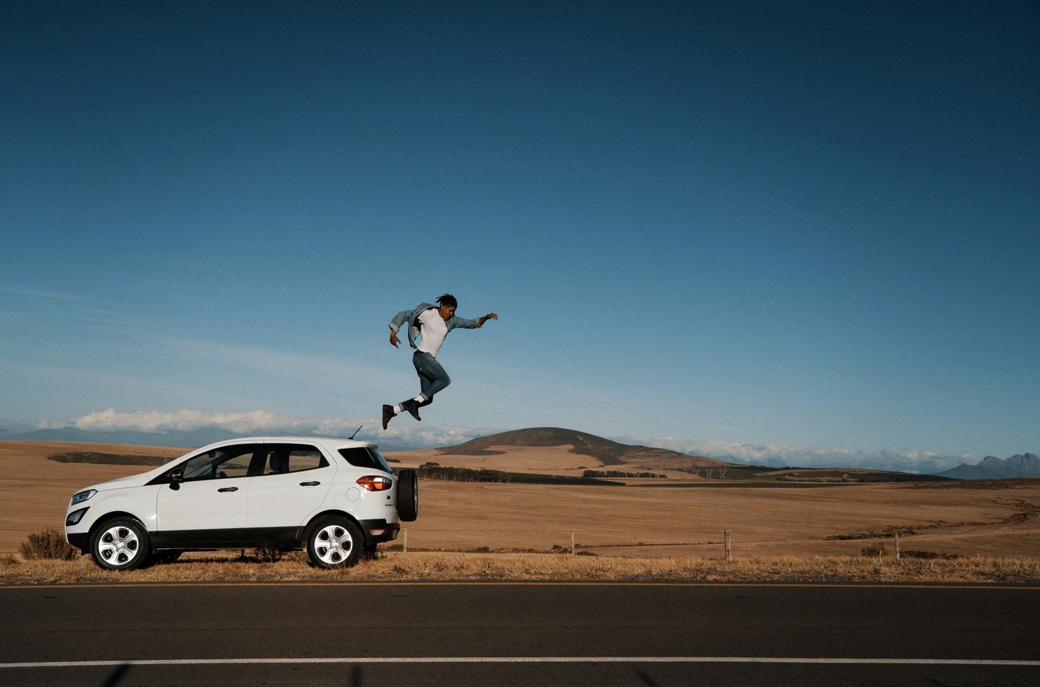 A MAN JUMPING FROM THE ROOF OF A WHITE FORD SUV IN AN OPEN LANDSCAPE OF HILLS.