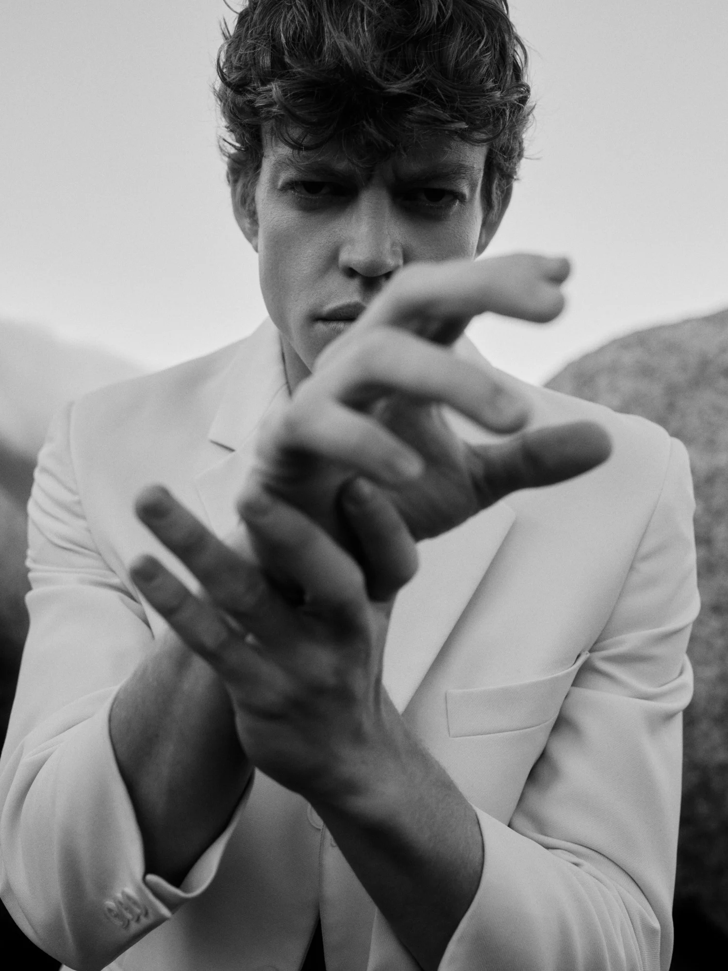 A MAN WITH CURLY HAIR AND A SERIOUS EXPRESSION, WEARING A LIGHT-COLORED BLAZER, IS REACHING OUT TOWARDS THE CAMERA WITH HIS HANDS IN THE FOREGROUND, IN A BLACK AND WHITE PHOTOGRAPH.
