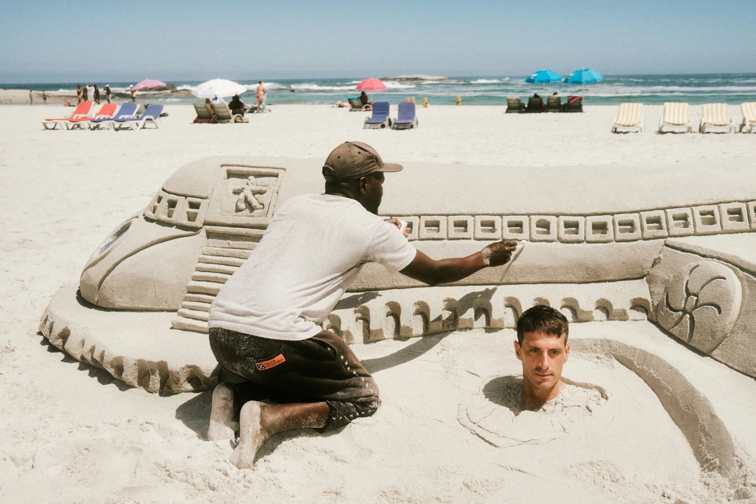 A MAN BURIED IN A SAND SCULPTURE OF A CLASSIC CAR, WITH BEACH UMBRELLAS AND THE OCEAN BEHIND.