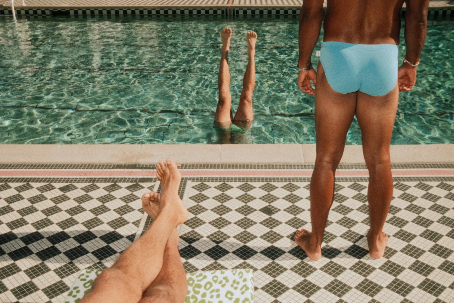 PEOPLE RELAXING POOLSIDE, WITH TWO PEOPLE FLOATING IN THE POOL AND ONE PERSON STANDING AT THE EDGE OF THE POOL.