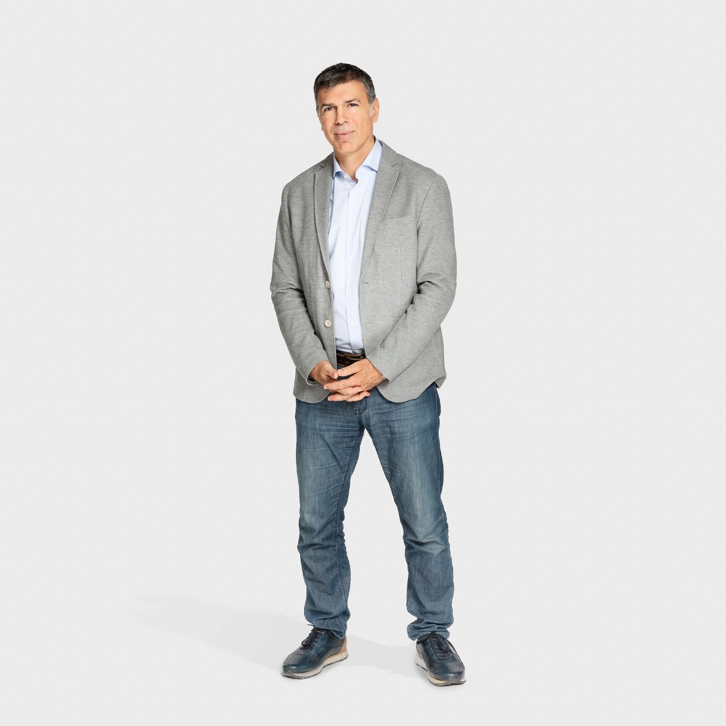 Full-length studio portrait of a man in a gray blazer, light blue shirt, and jeans, standing with his hands clasped against a pale background.