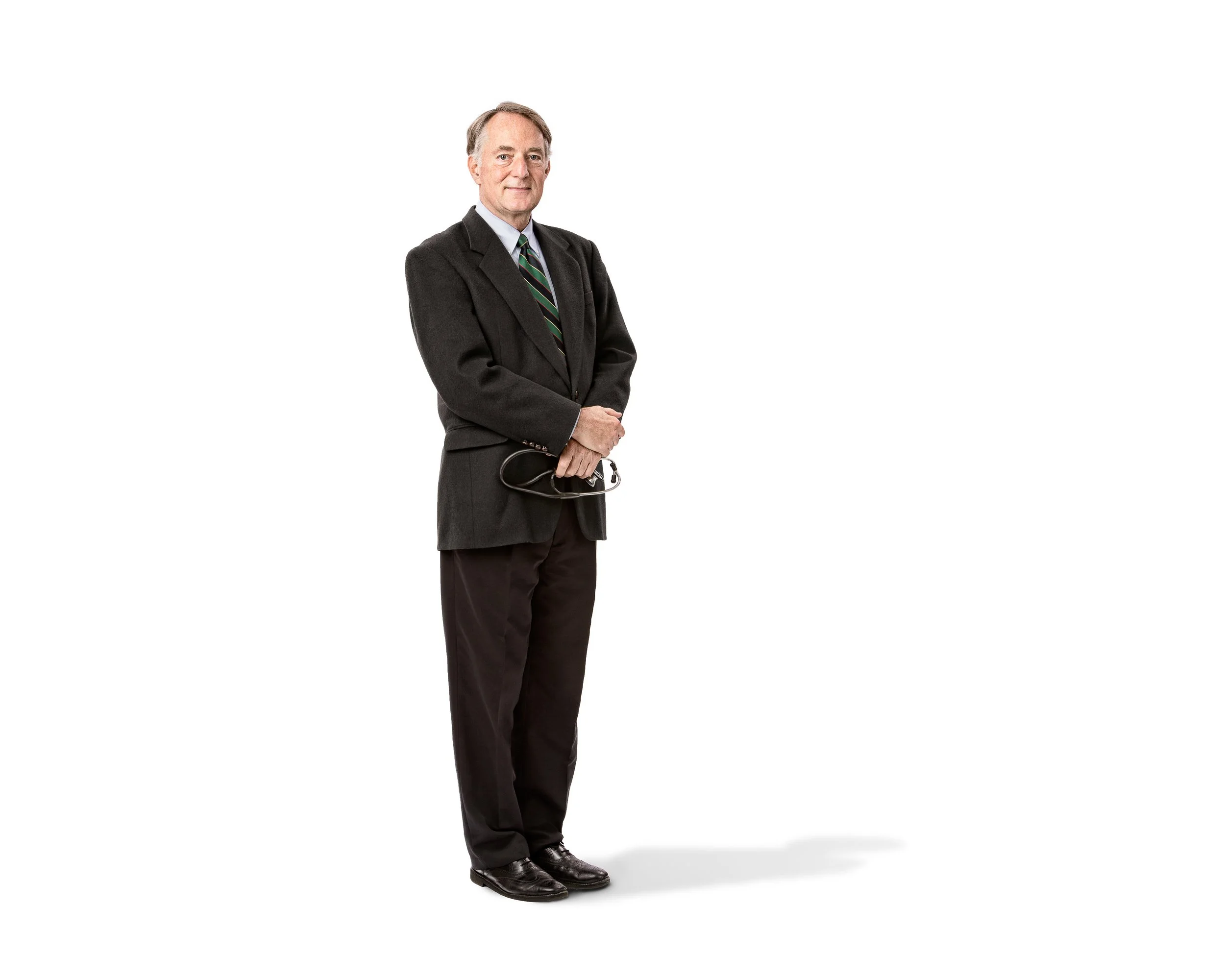 A full-length professional portrait of an older man wearing a dark suit and holding his glasses against a white background.