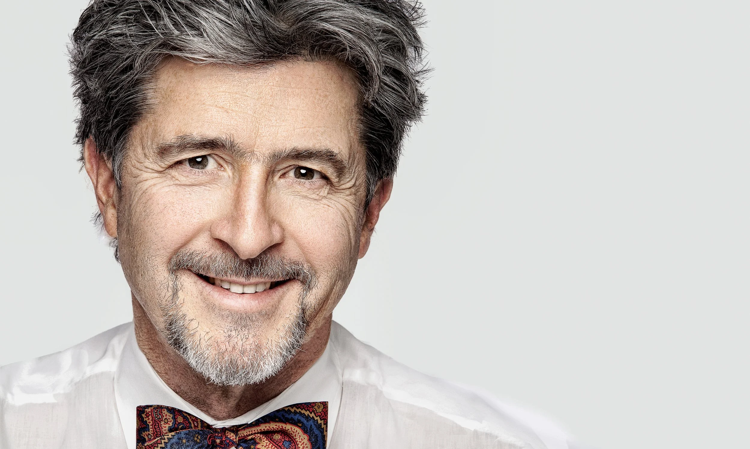 A close-up professional portrait of a smiling mature man wearing a white shirt and patterned bow tie.