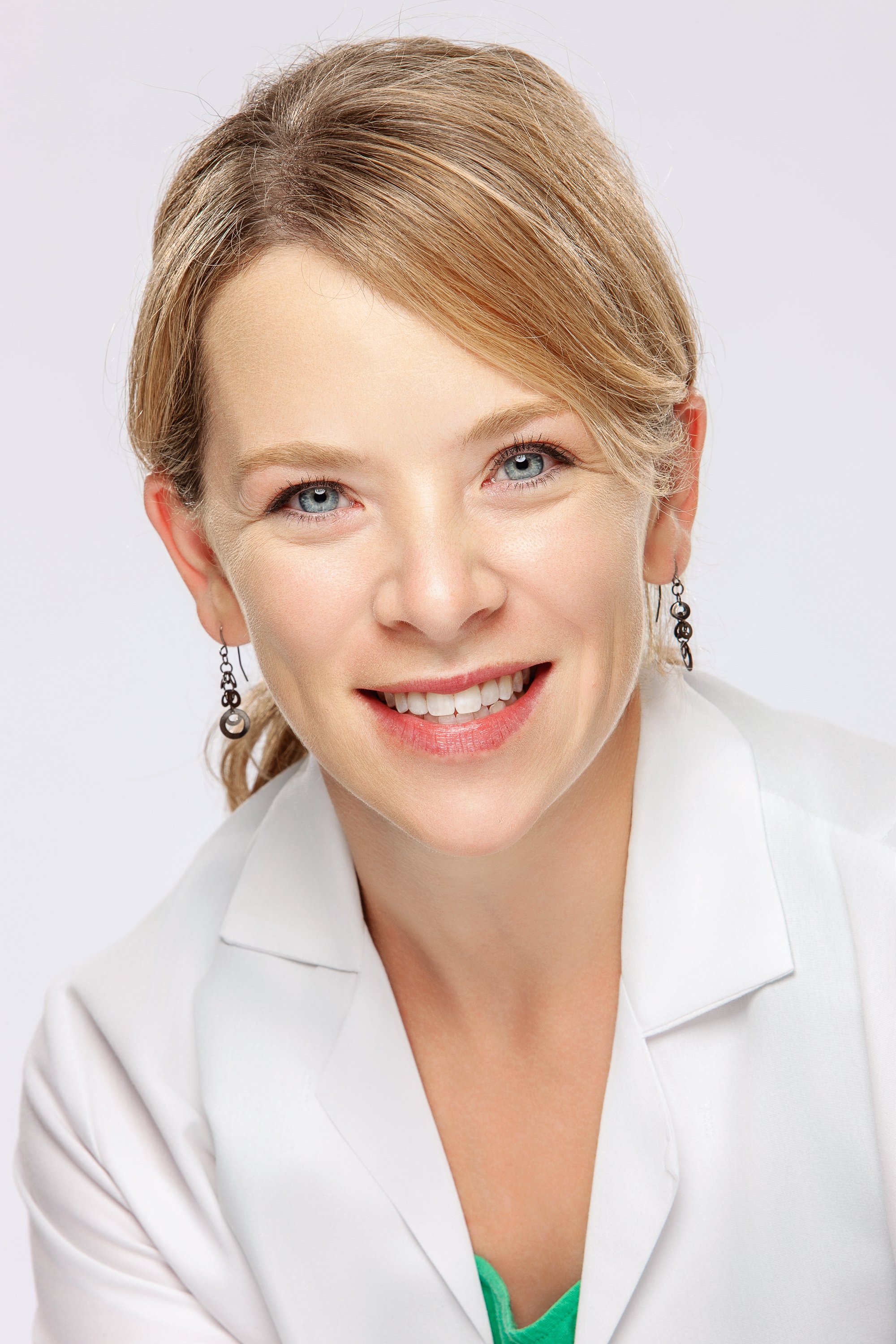 Headshot of a smiling woman with blonde hair and dangling earrings, wearing a white coat over a green top against a pale background.