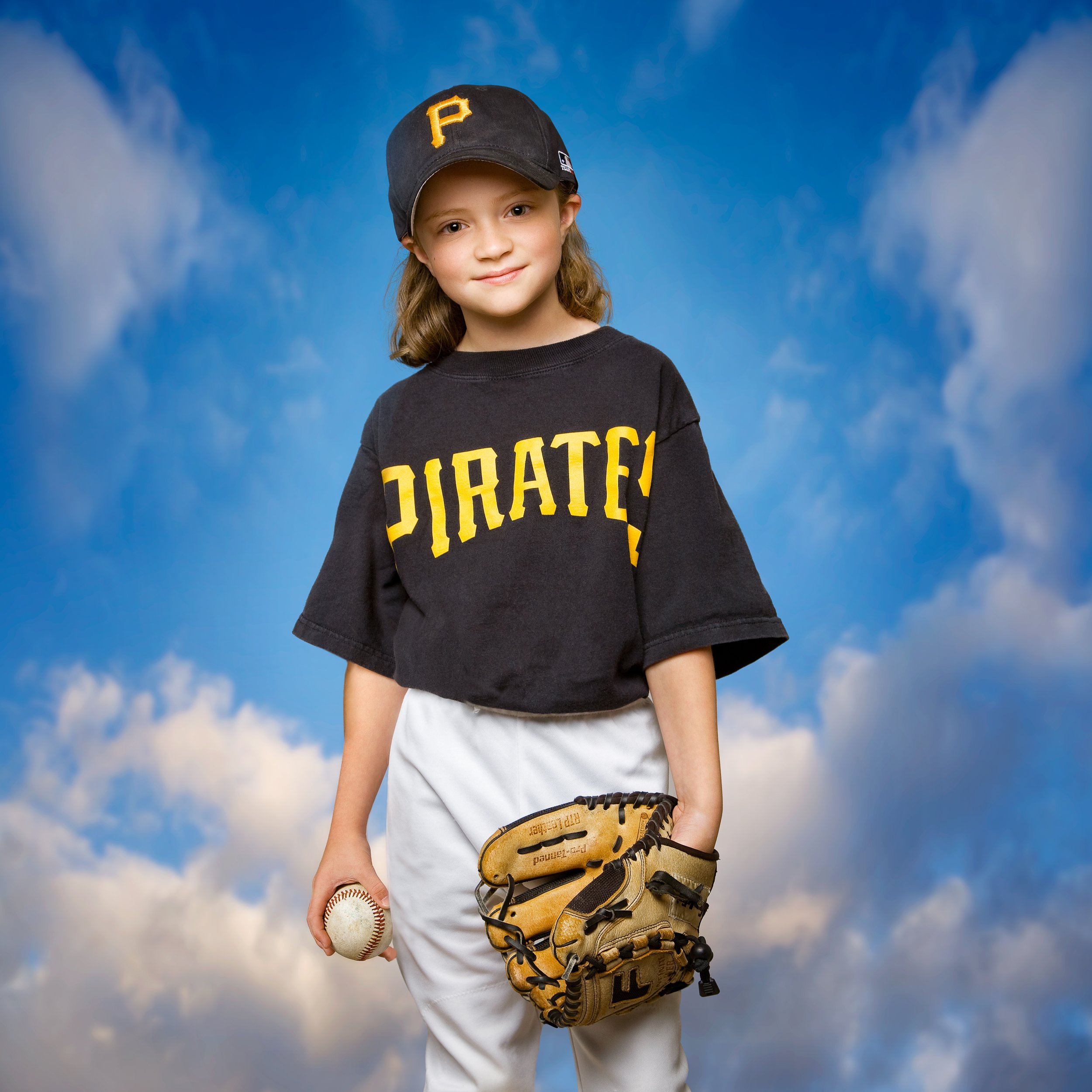 Studio photograph of a child in a Pirates baseball uniform and cap, holding a ball and glove against a blue sky and cloud backdrop.