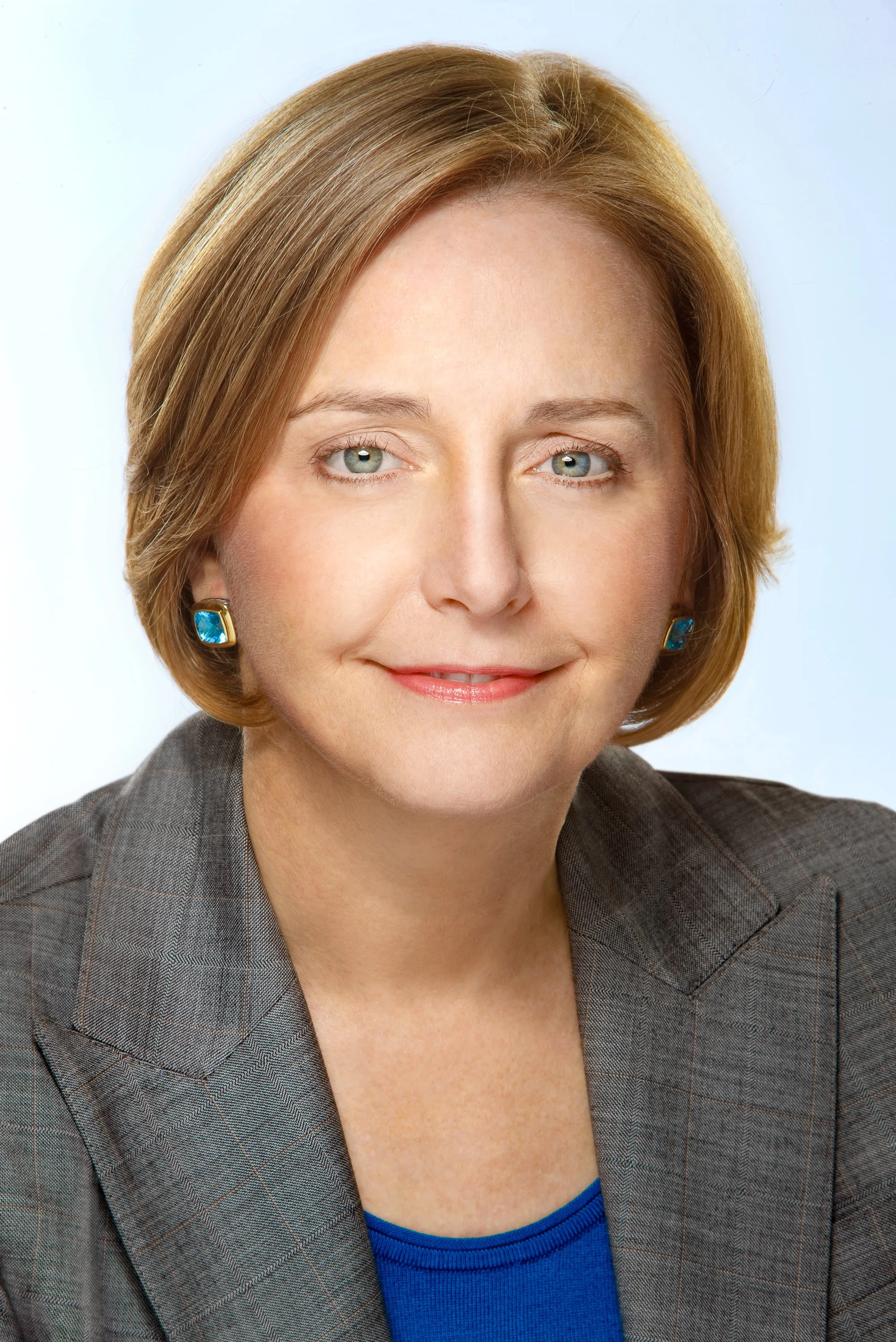 Headshot of a woman with short auburn hair wearing a gray blazer, blue top, and turquoise earrings against a pale blue background.