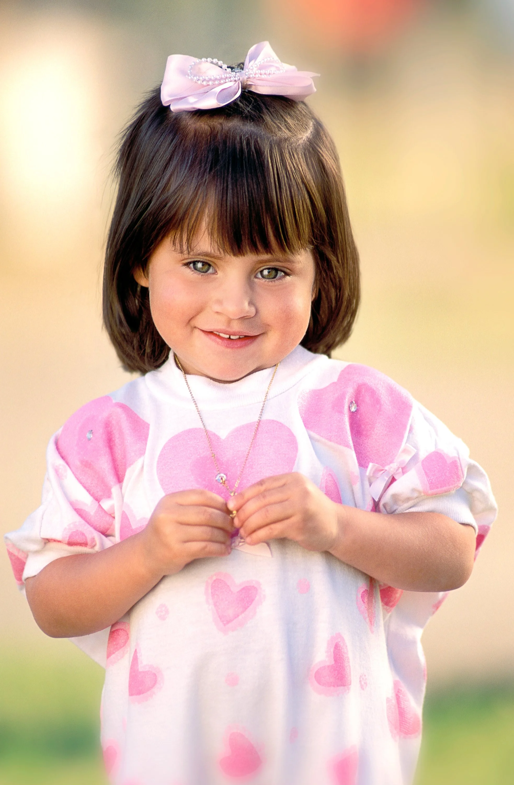 A young girl wearing a pink dress and matching hair bow photographed against a softly blurred outdoor background.