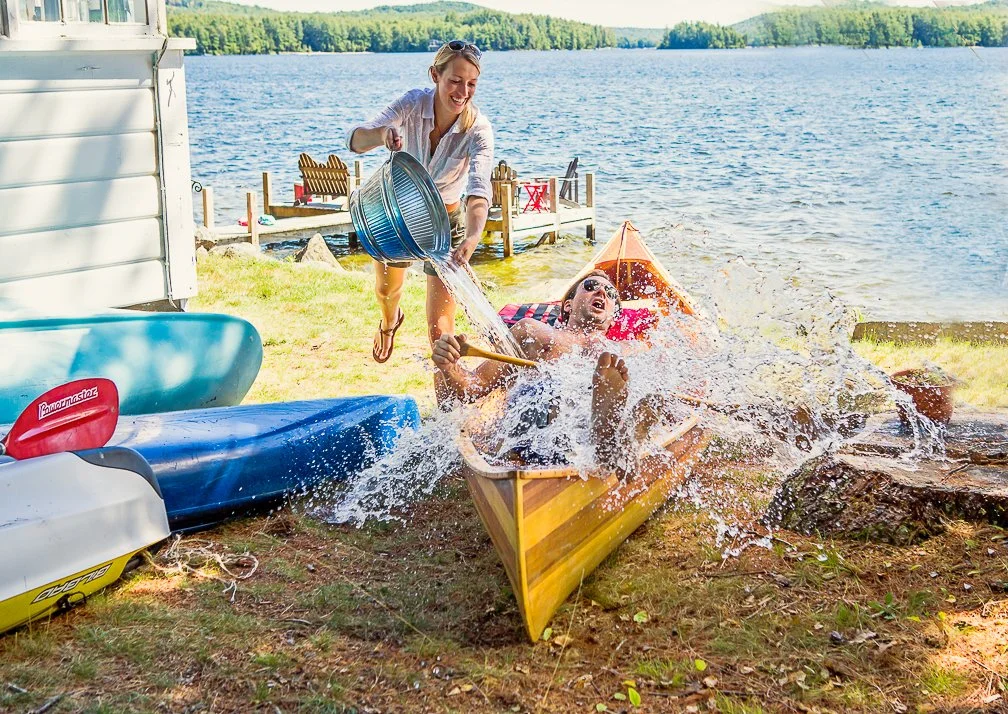 A young couple at Lake Winnipesaukee. An energetic lakeside moment as a woman playfully dumps a bucket of water onto a friend lounging in a canoe, creating a splash of laughter and surprise. Set against the scenic New Hampshire lake backdrop with ka