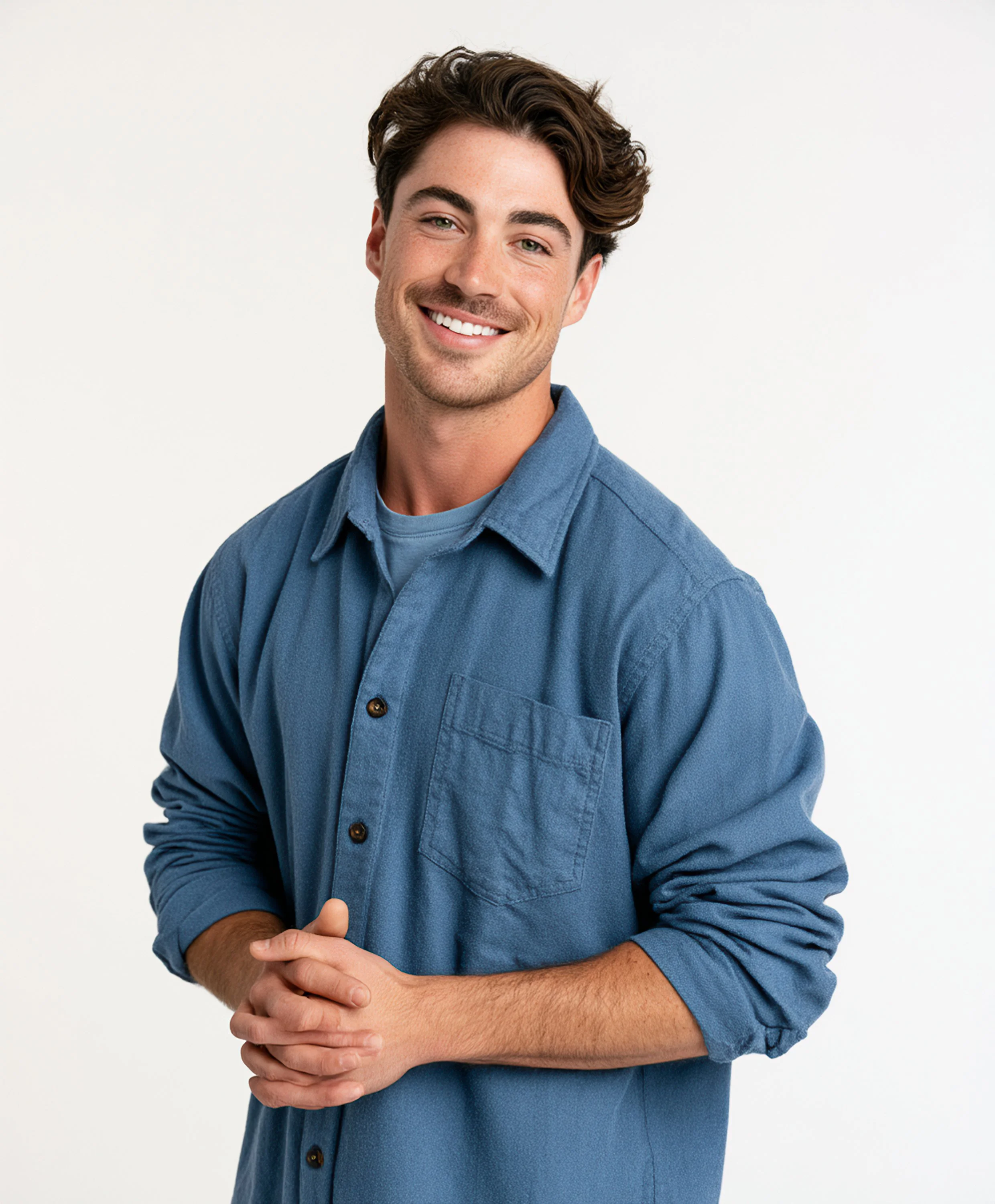 Smiling man with wavy dark hair wearing a blue overshirt with rolled sleeves, his hands loosely clasped against a white background.