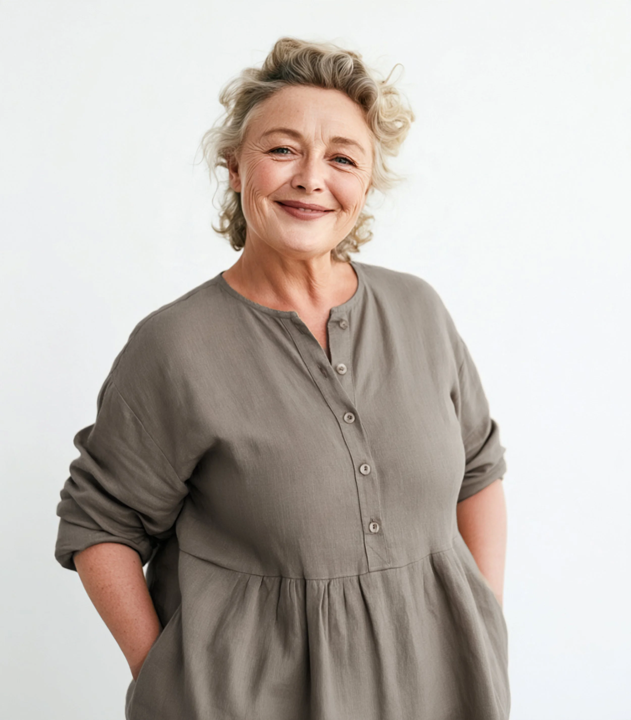 Relaxed portrait of a smiling woman with short gray curls, wearing a loose gray dress with her hands in its pockets against a white background.