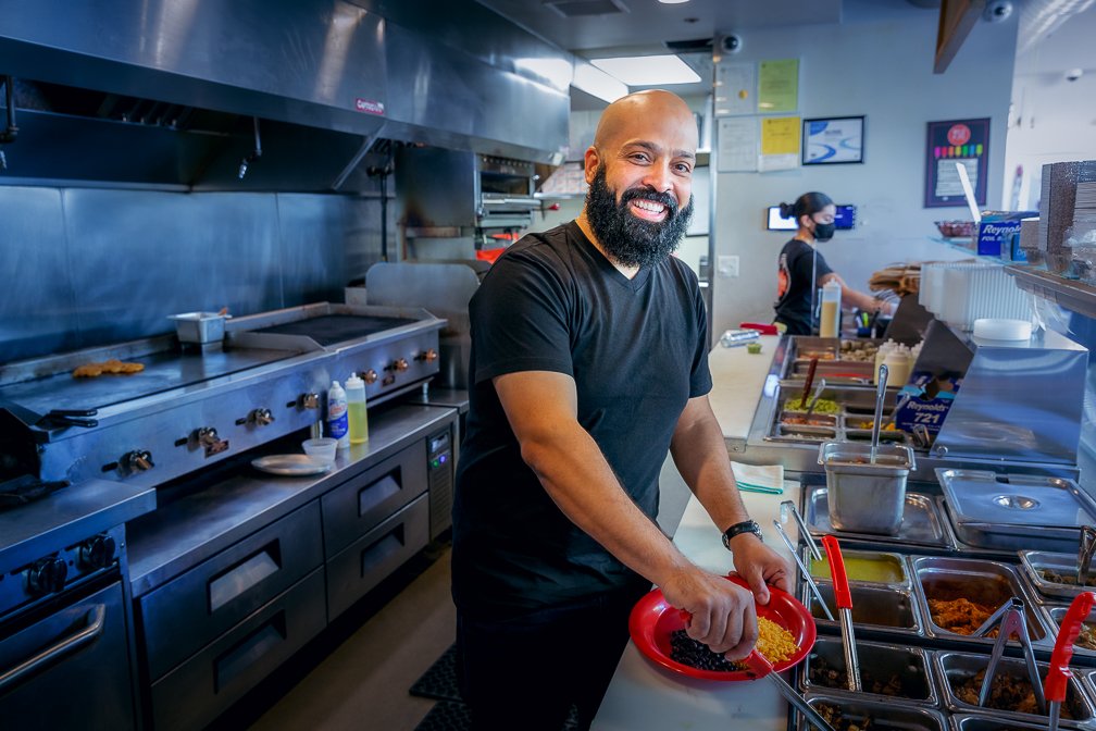 The proud minority owner of a brand-new sub shop smiling happily as he preps a fast-food order.