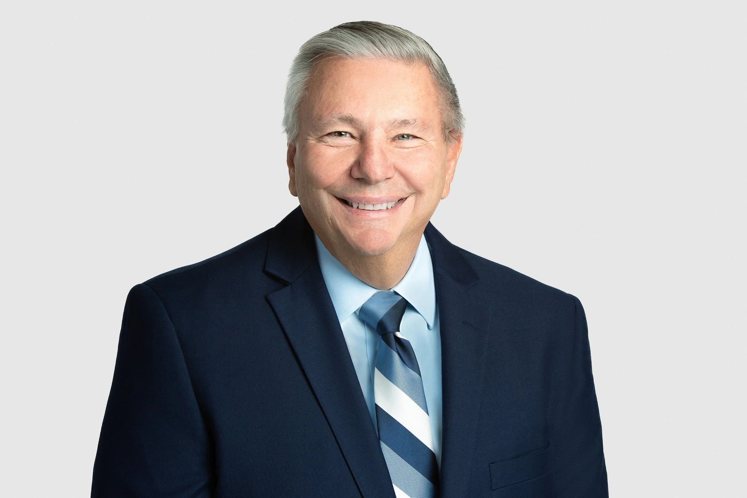 A professional portrait of a smiling older man wearing a navy-blue suit and striped tie against a light gray background.