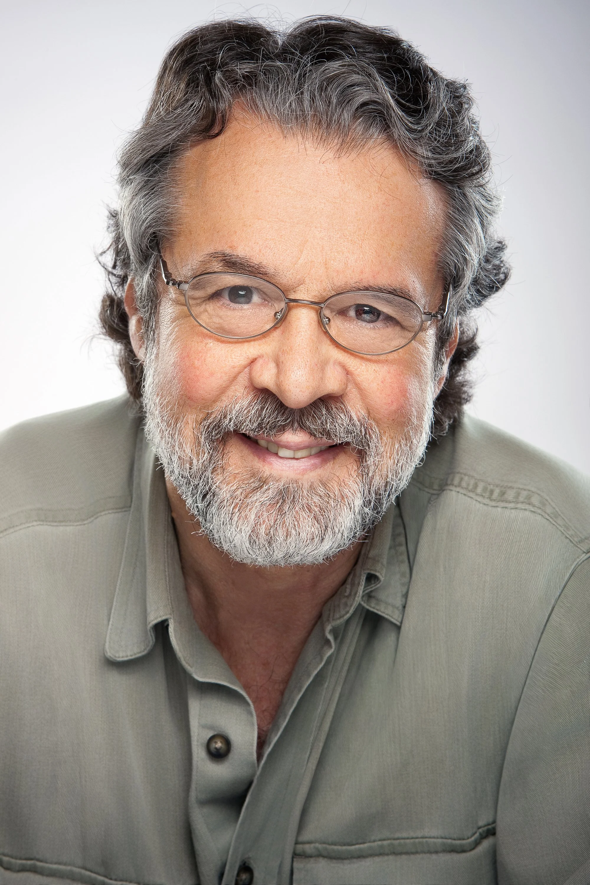 A relaxed portrait of an older man with gray hair, glasses, and a full beard wearing a casual gray shirt.