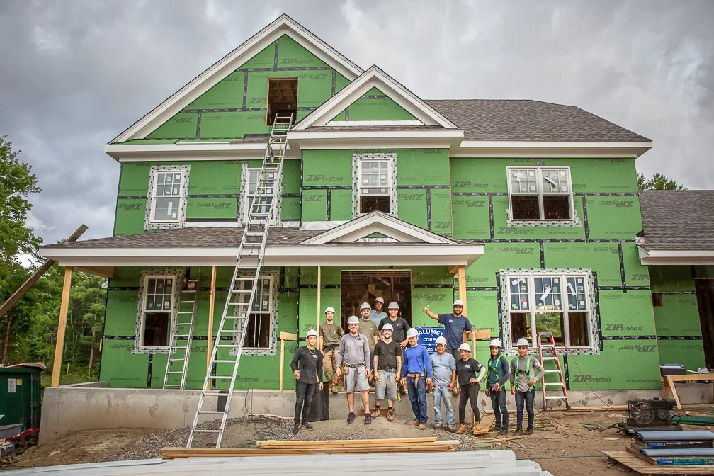 A full crew of house construction workers proudly posing in front of the nearly completed house they built.