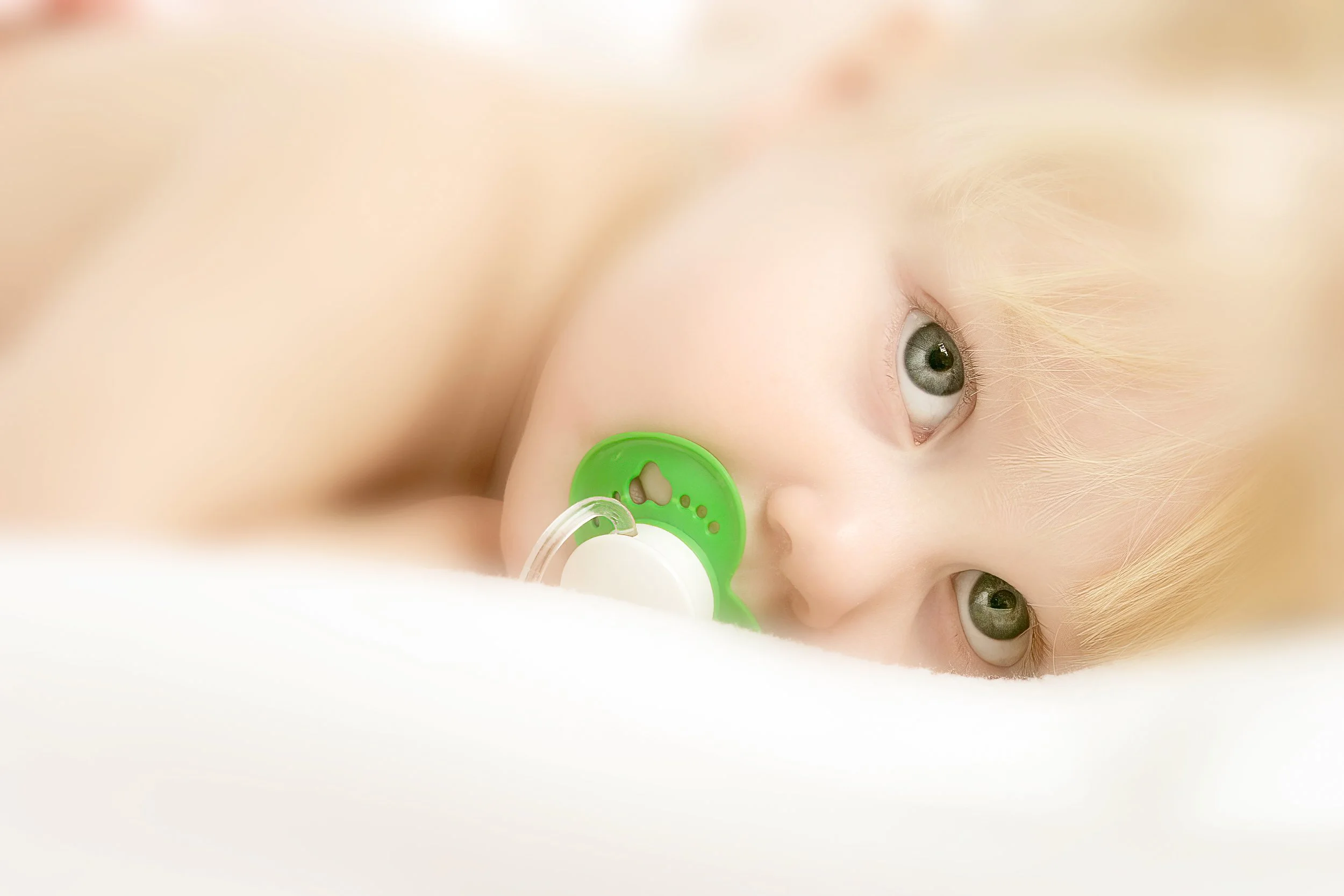 A fair-haired baby resting peacefully with a green pacifier against soft white bedding.