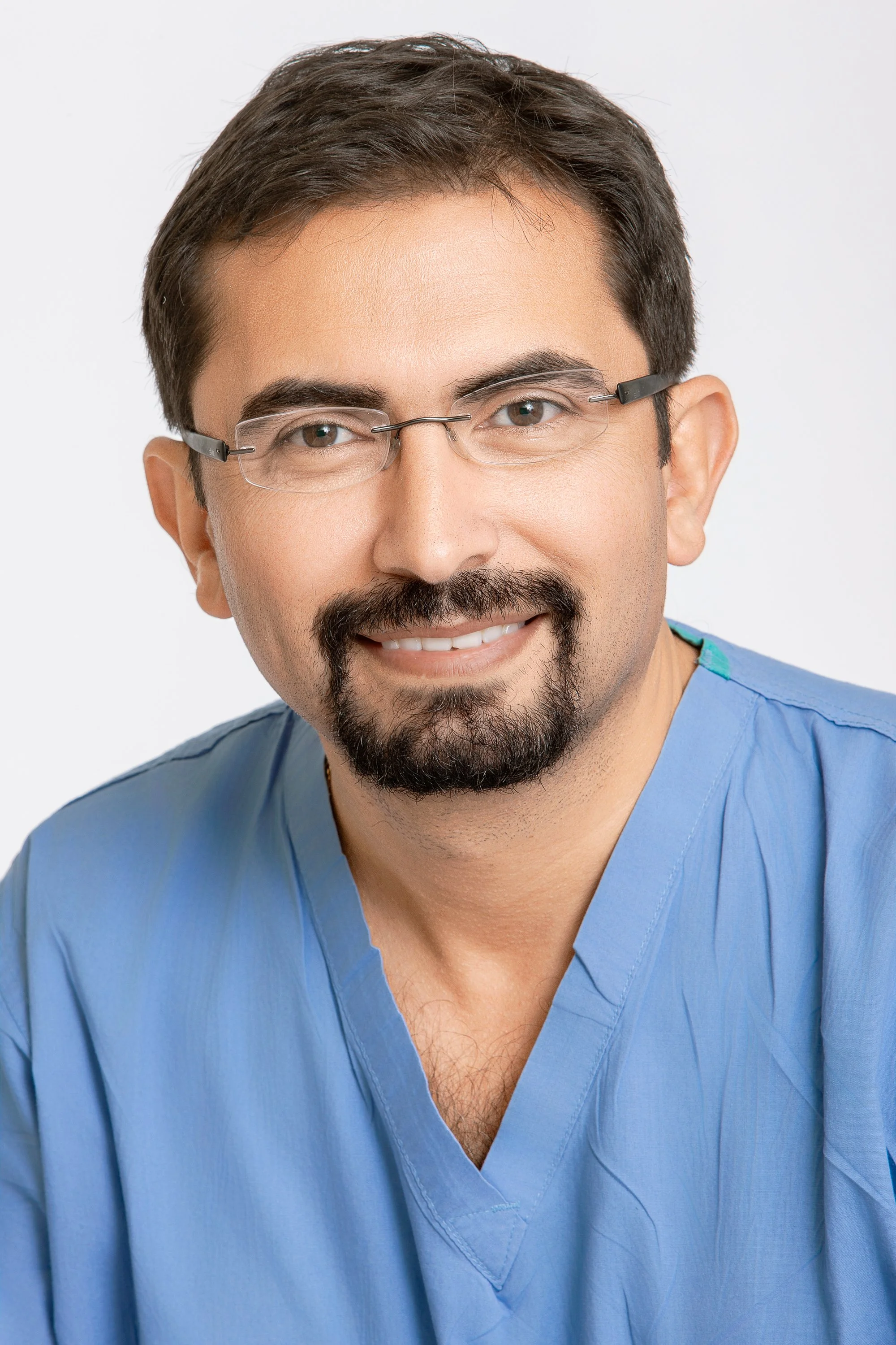 Portrait of a smiling man with glasses, dark hair, and a neatly trimmed beard, wearing blue medical scrubs against a white background.