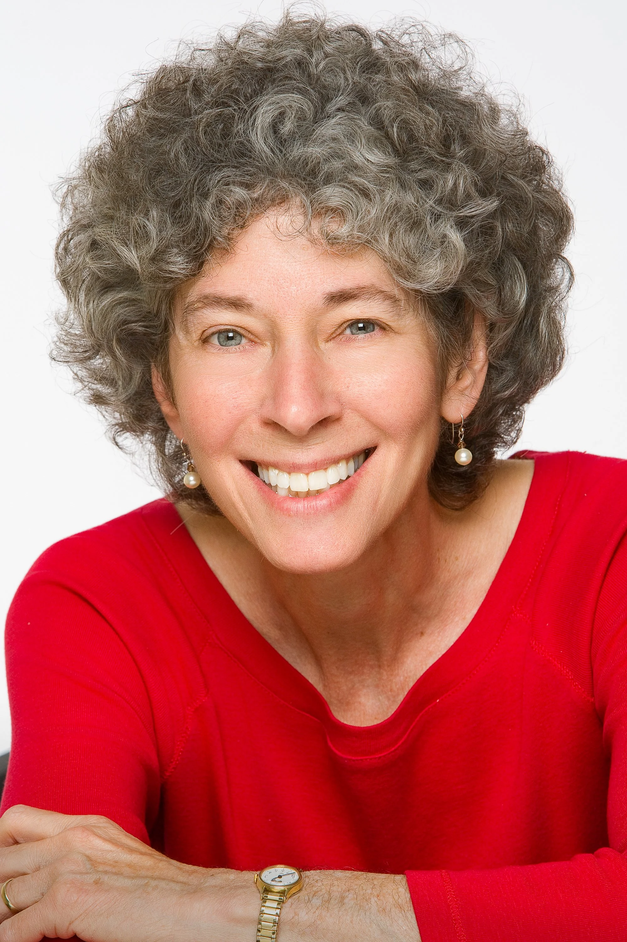 Smiling woman with curly gray hair wearing a bright red top and pearl earrings, leaning forward with her arms folded against a white background.