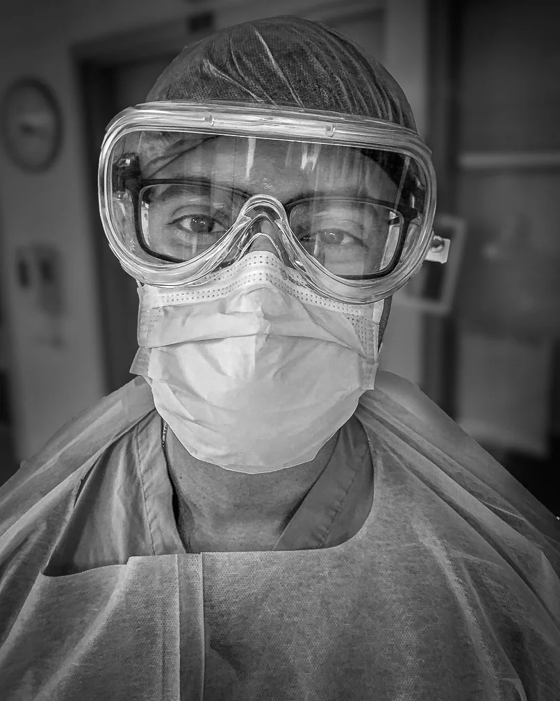 An extreme close-up journalistic photograph of a young doctor with total face protection, showing tired yet optimistic eyes.