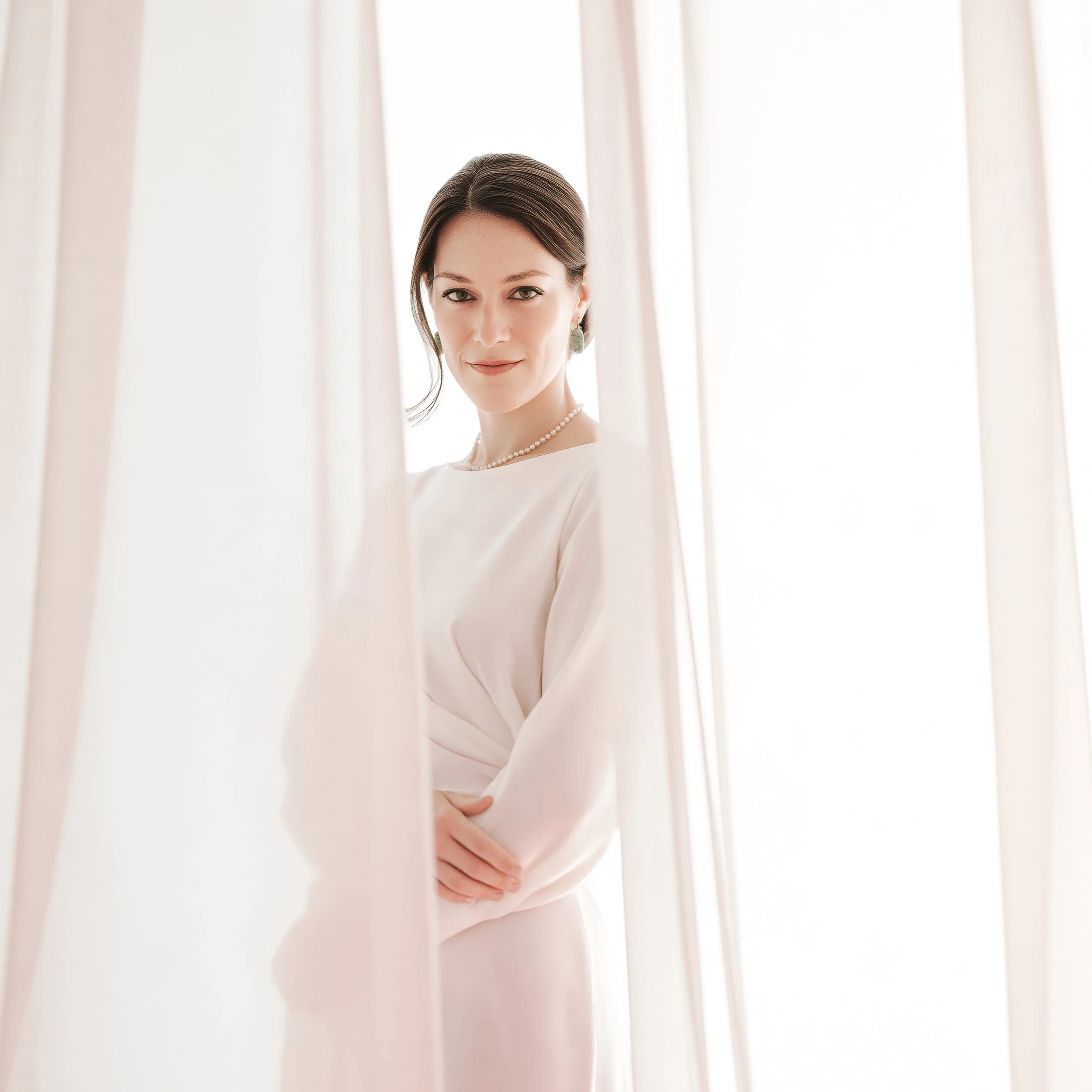 Woman in a white dress and pearl necklace, framed by sheer curtains in soft, bright light.