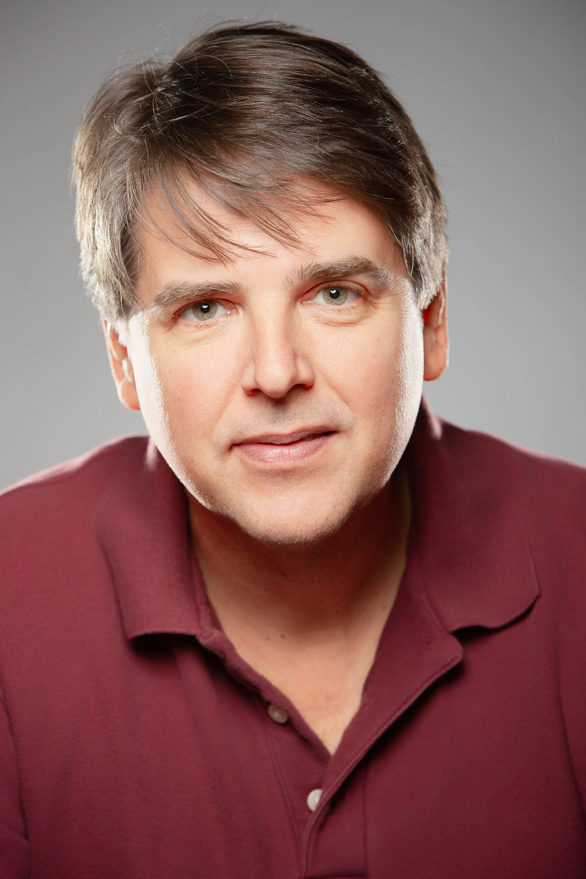 Close-up portrait of a man with brown hair wearing a burgundy polo shirt against a gray background.