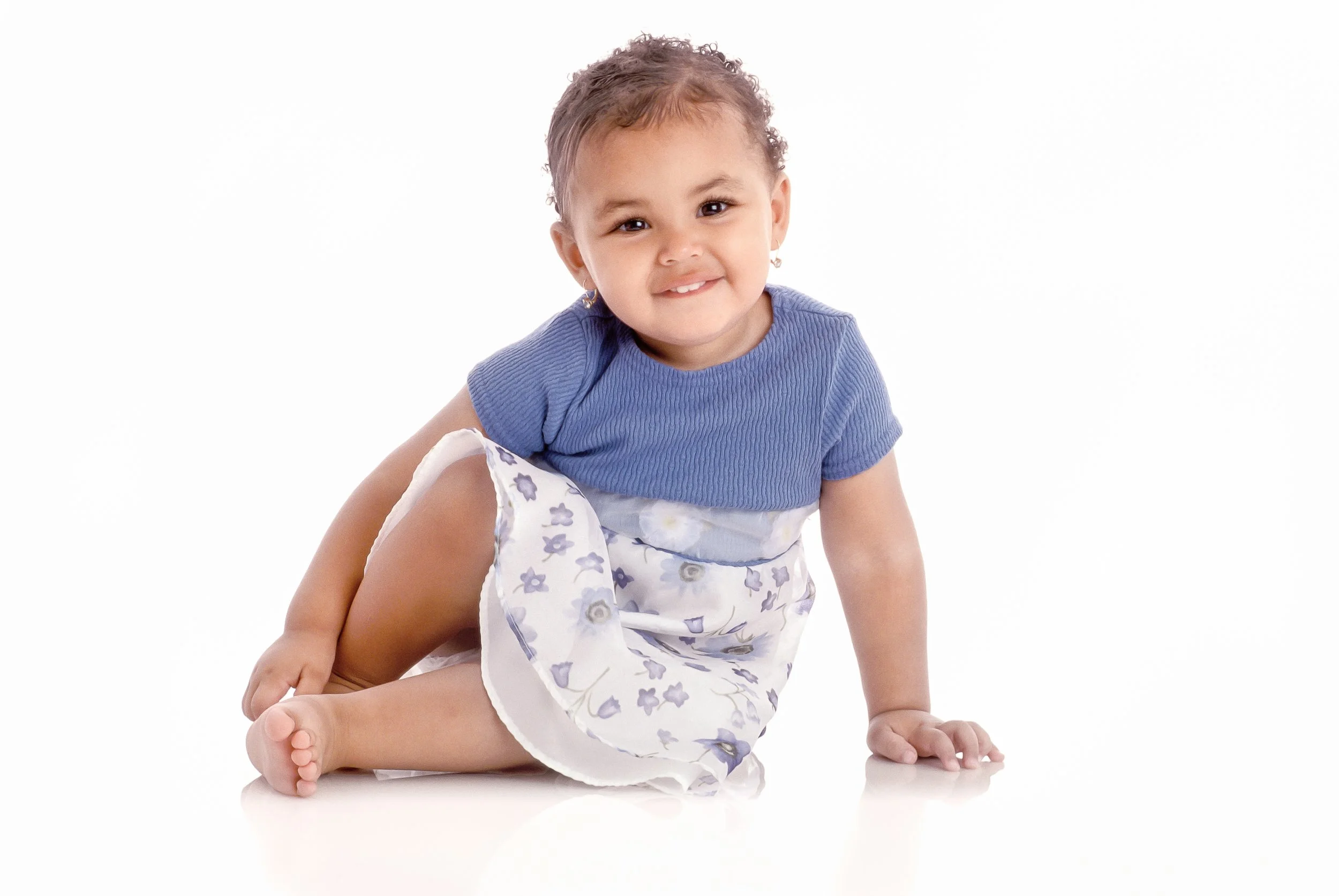 A smiling toddler seated comfortably on the floor against a bright white studio background.