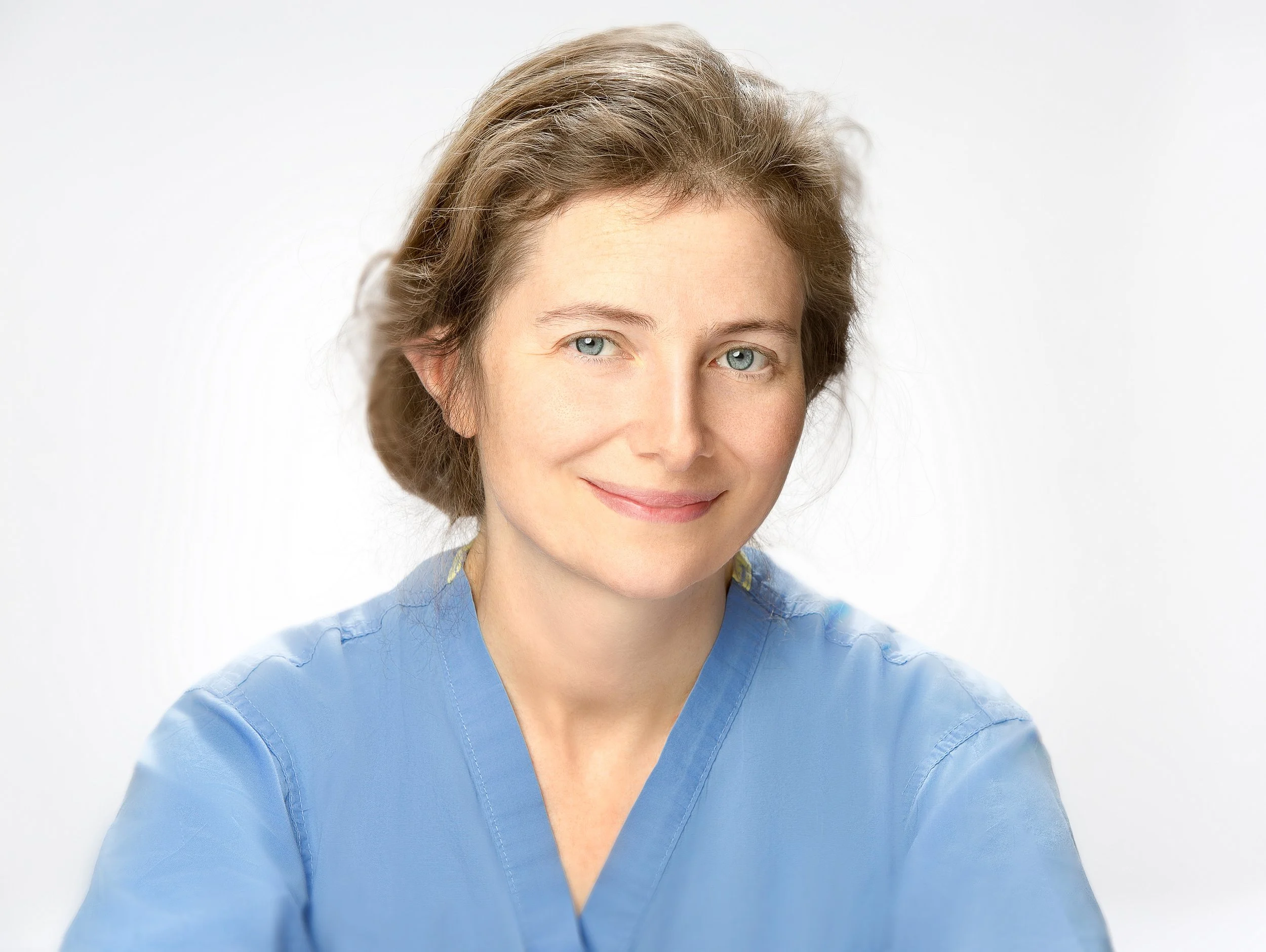 Close-up portrait of a smiling doctor with brown hair wearing blue medical scrubs against a white background.