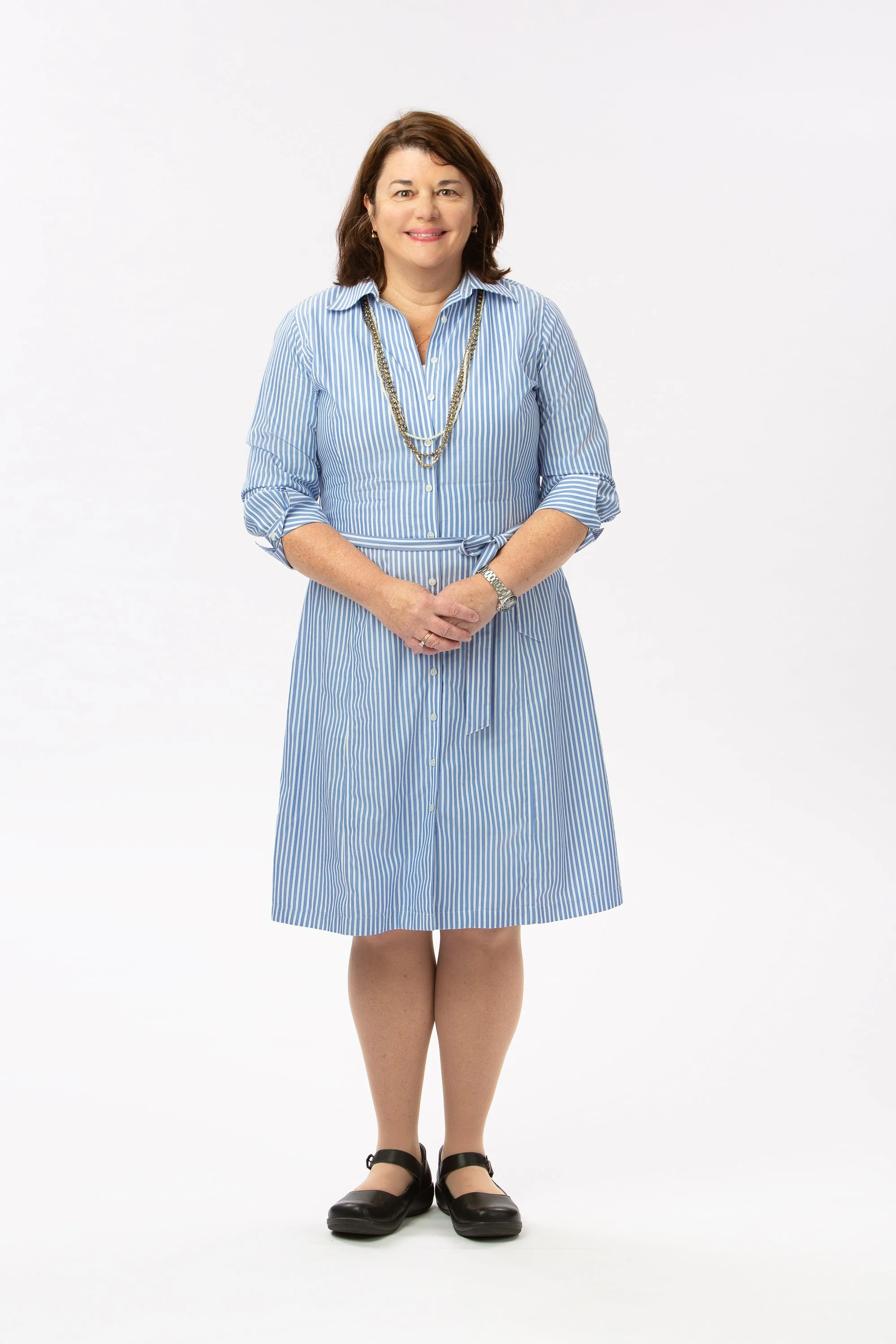 Full-length studio portrait of a woman in a blue striped dress, long necklace, and black shoes, standing with her hands clasped against a white background.