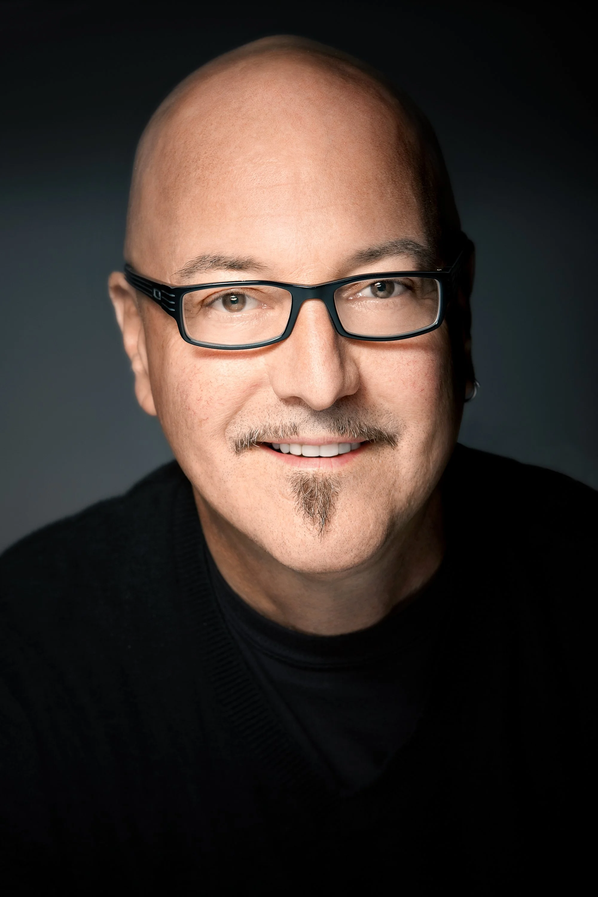 Smiling bald man with black rectangular glasses, a mustache, and a small goatee, wearing black against a dark background.