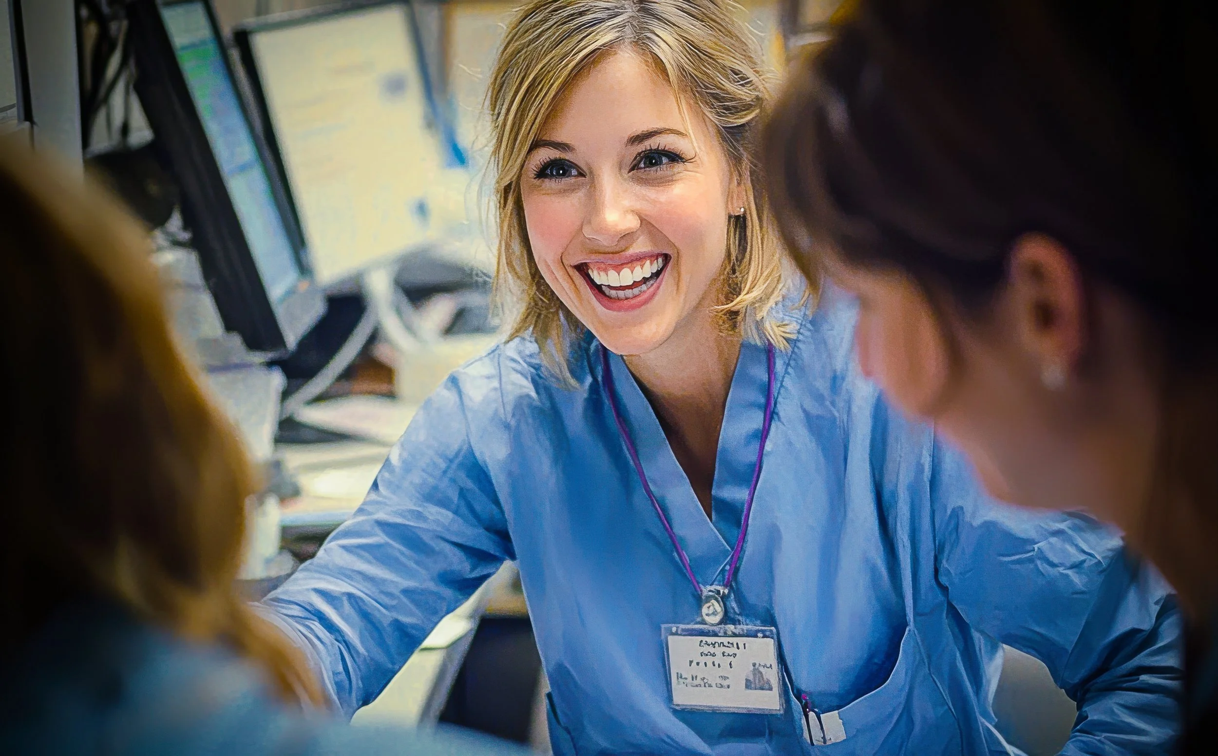 A smiling healthcare professional in blue scrubs talks with colleagues at a busy hospital workstation.