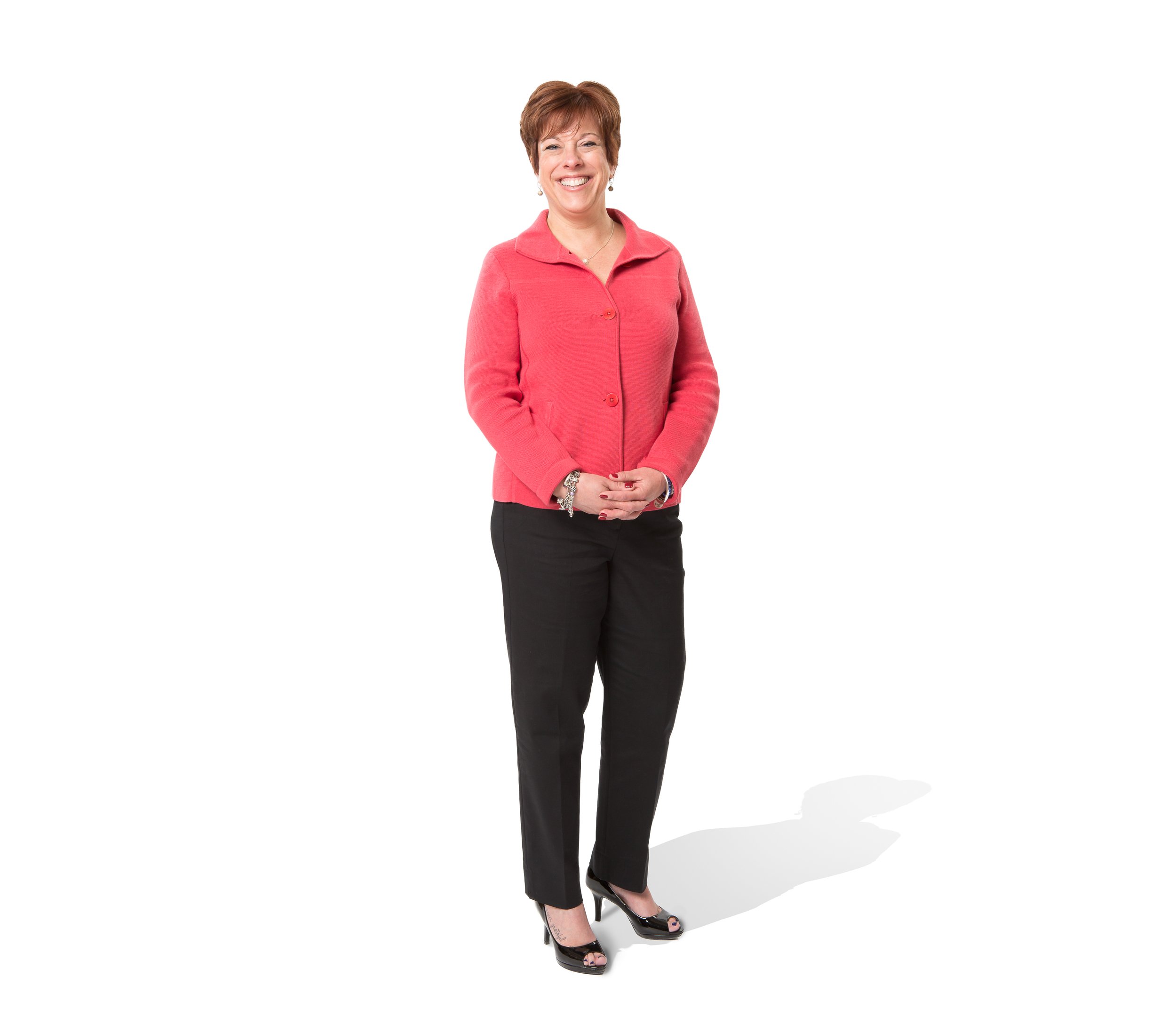 A full-length portrait of a smiling mature woman wearing a coral-colored sweater and black trousers.
