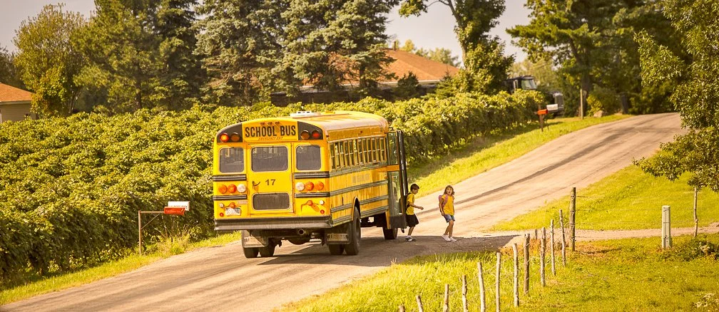 Two school children wearing summer shorts getting off the big yellow school bus in a rural country setting.