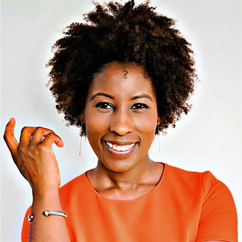 Smiling person with curly hair wearing an orange shirt, holding a hand near their face.