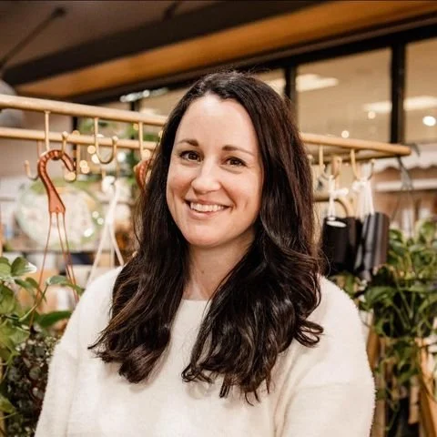 Smiling person with long dark hair wearing a white sweater, standing in front of indoor plants and hanging decor in a well-lit room.