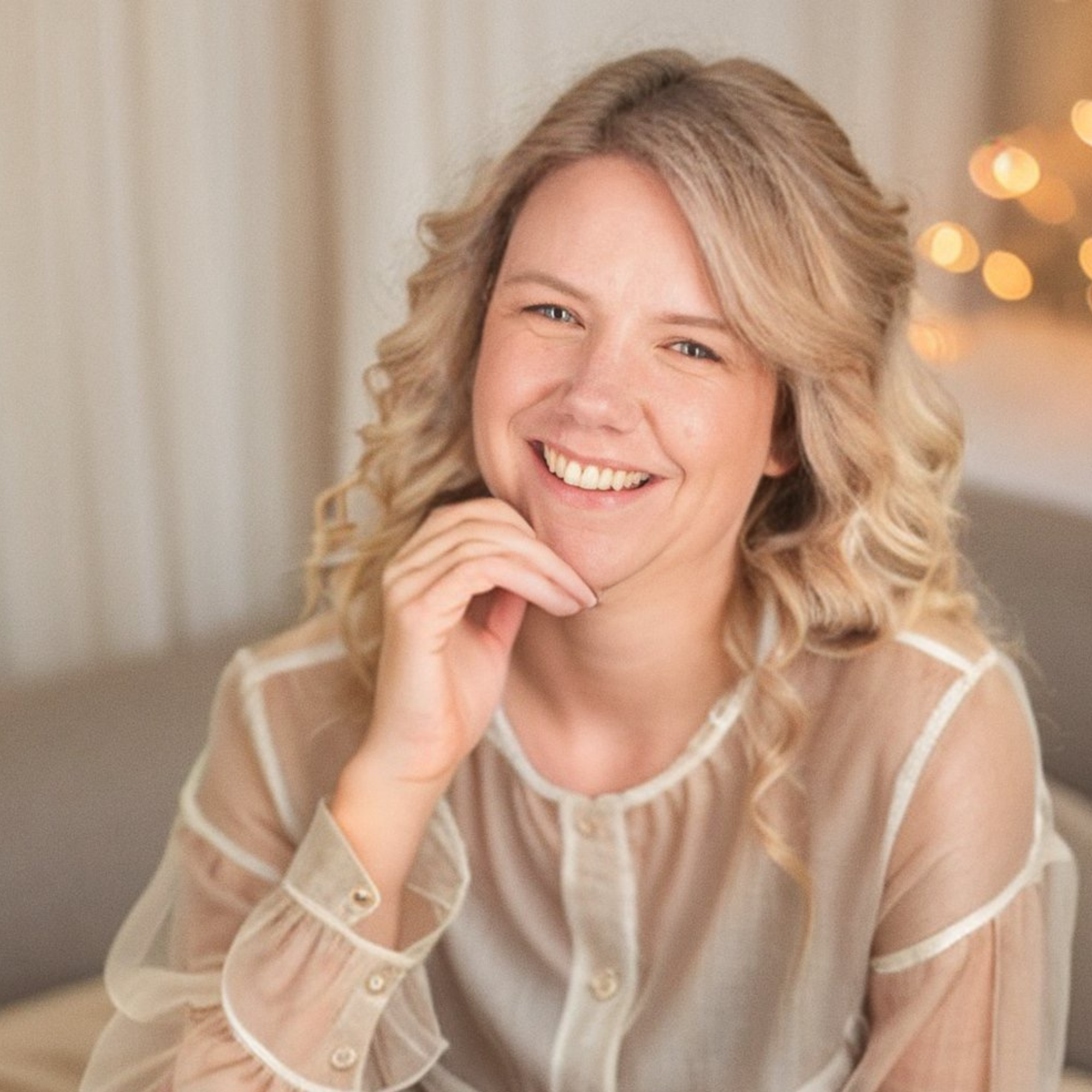 Close-up of a woman with curly blonde hair smiling, wearing a beige sheer blouse with buttons, in warm indoor lighting with blurred bokeh lights in the background.