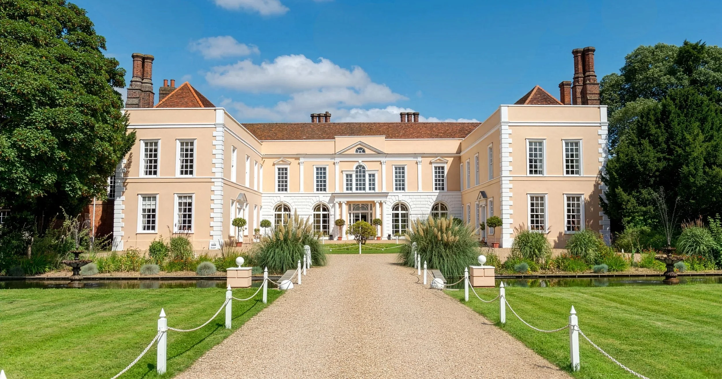 Large beige mansion with white accents, multiple windows, and a central entrance, surrounded by a well-maintained garden with trees, shrubs, and a gravel pathway leading to the front door, under a blue sky with some clouds.